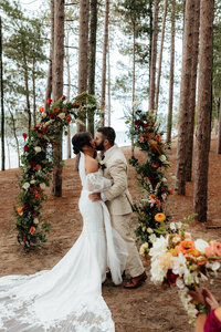 bride and groom kissing in tall pine trees with a floral arch as the backdrop