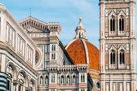 The image shows the Florence Cathedral, featuring its ornate façade with intricate designs and a prominent red-tiled dome. The adjacent bell tower, adorned with decorative patterns, stands tall against a clear blue sky.