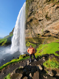 Brandi standing under a waterfall with a green lush background and the sun shining