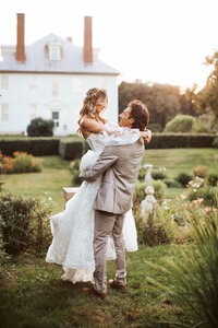 A groom picks up his bride and spins her around in a garden of a historic venue.