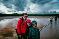 Gina Cooperman with her two children, wearing vibrant cold weather gear, in front of a geothermal pool under stormy skies.