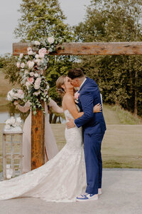 Bride and groom kissing under wooden arch with pink and white floral arrangement.