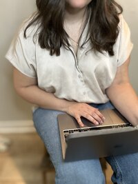 Close-up of a woman’s hands typing on a silver laptop, wearing a light blouse and blue jeans.