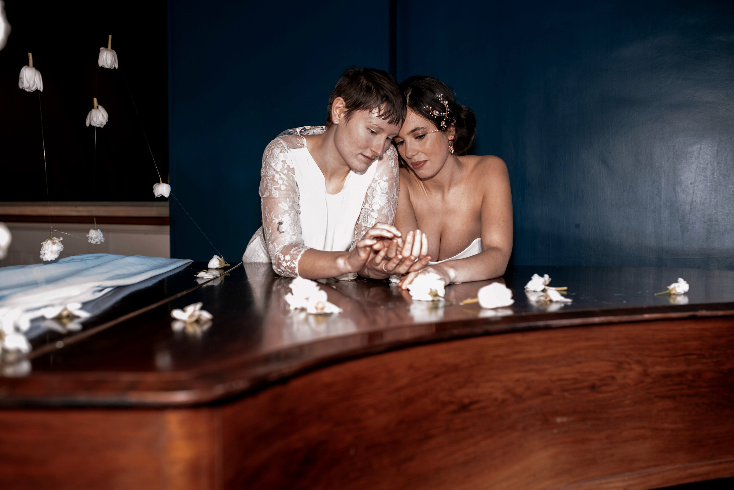 Two brides holding hands leaning on a wooden piano surrounded by hanging flowers