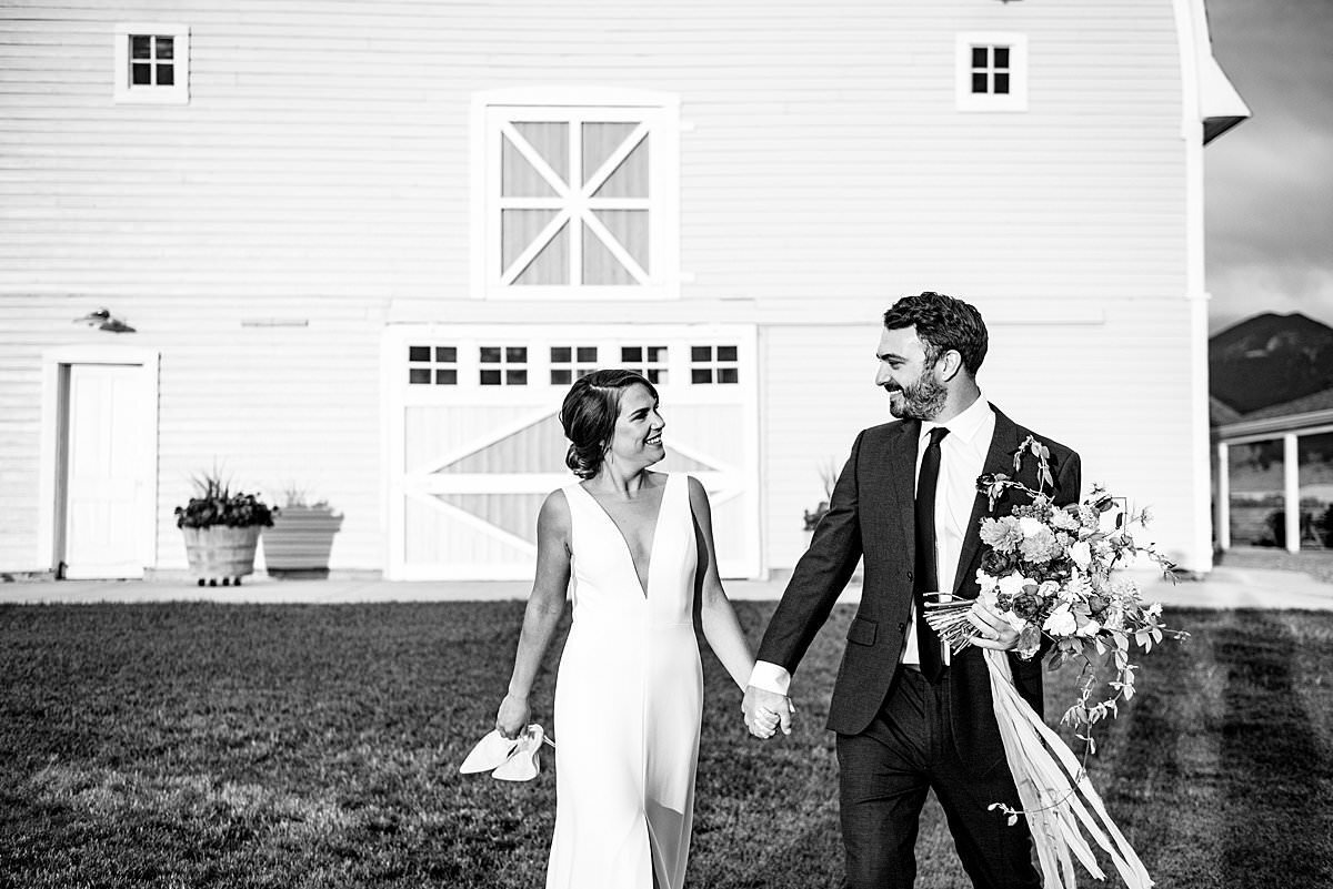 Black and white photo bride and groom walking together in front of big yellow barn in Bozeman, Montana