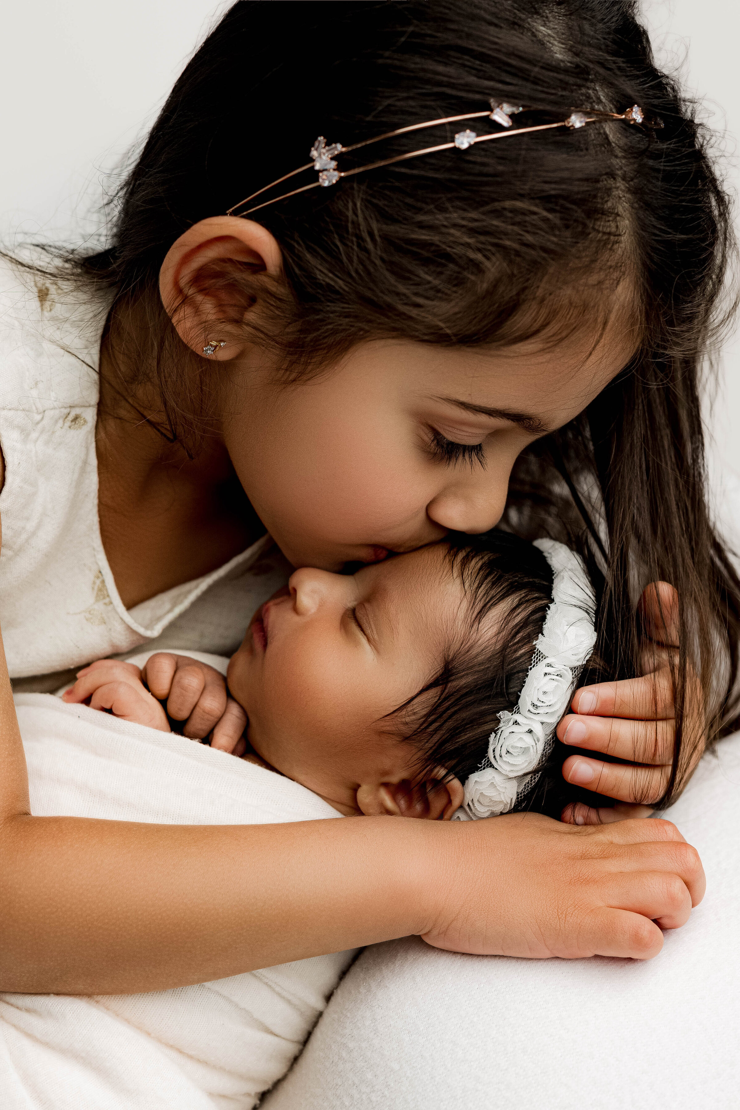 Big sister sweetly kissing her newborn sibling on the forehead, wrapped in white and nestled closely.