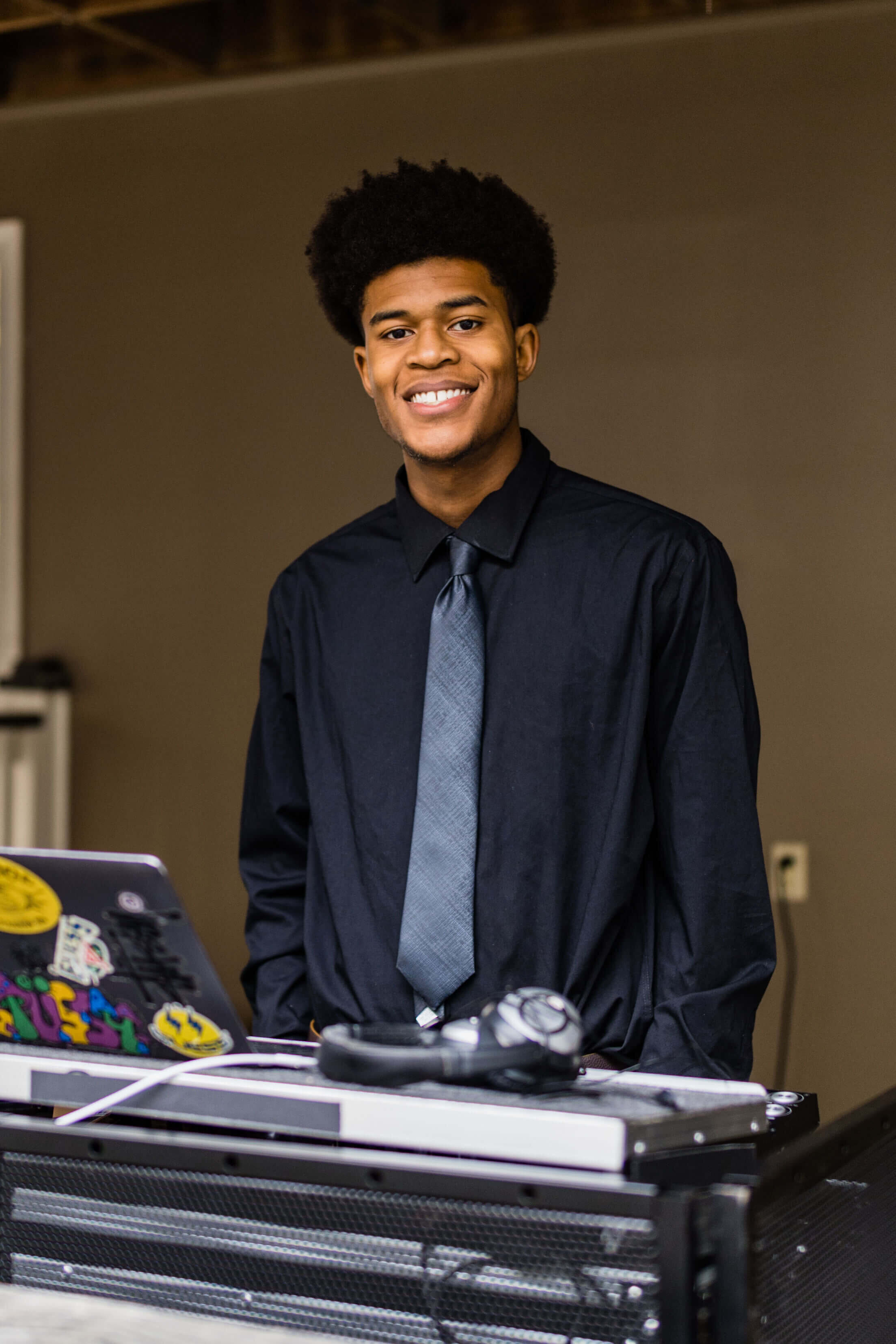 A young man with curly hair wearing a black shirt and gray tie smiles while standing behind a DJ booth with a laptop and headphones.