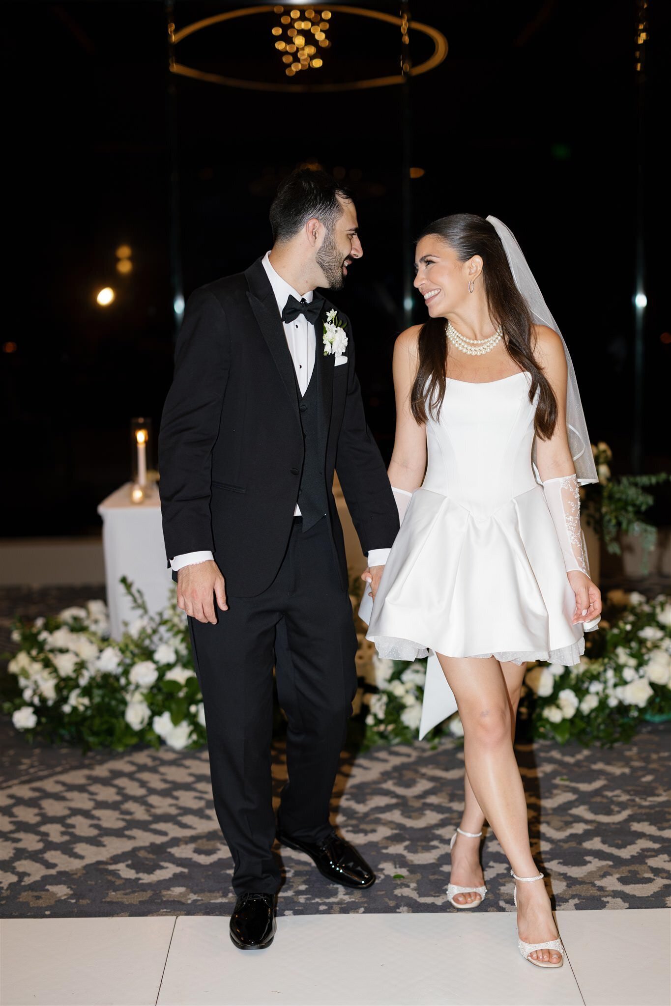 A newlywed couple holding hands and walking as they smile at each other 