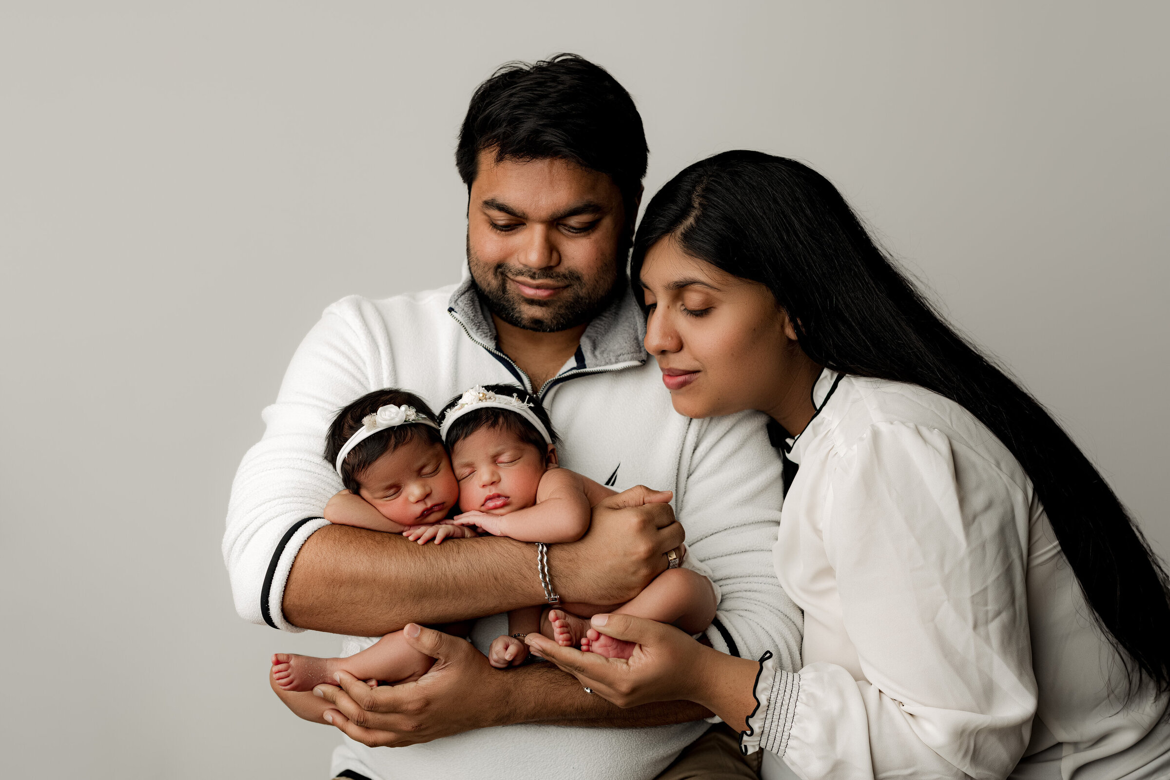 A couple lovingly holds their two sleeping newborn babies, captured beautifully by a Charlotte newborn photographer. The babies, dressed in white with matching headbands, are cradled gently by their parents, who gaze down fondly at them.