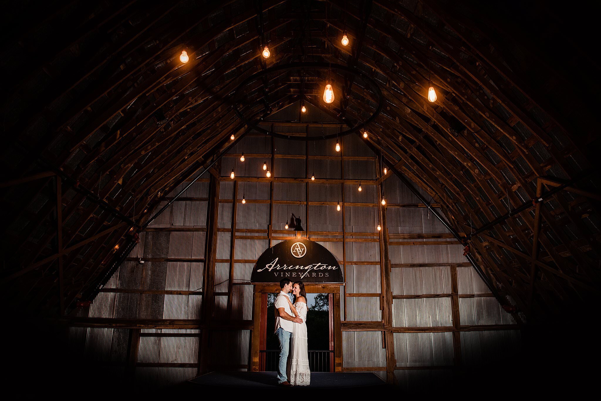 Engaged couple sharing a romantic slow dance on stage in the balcony of the big red barn at Arrington Vineyards, dramatic lighting on them with chandeliers overhead