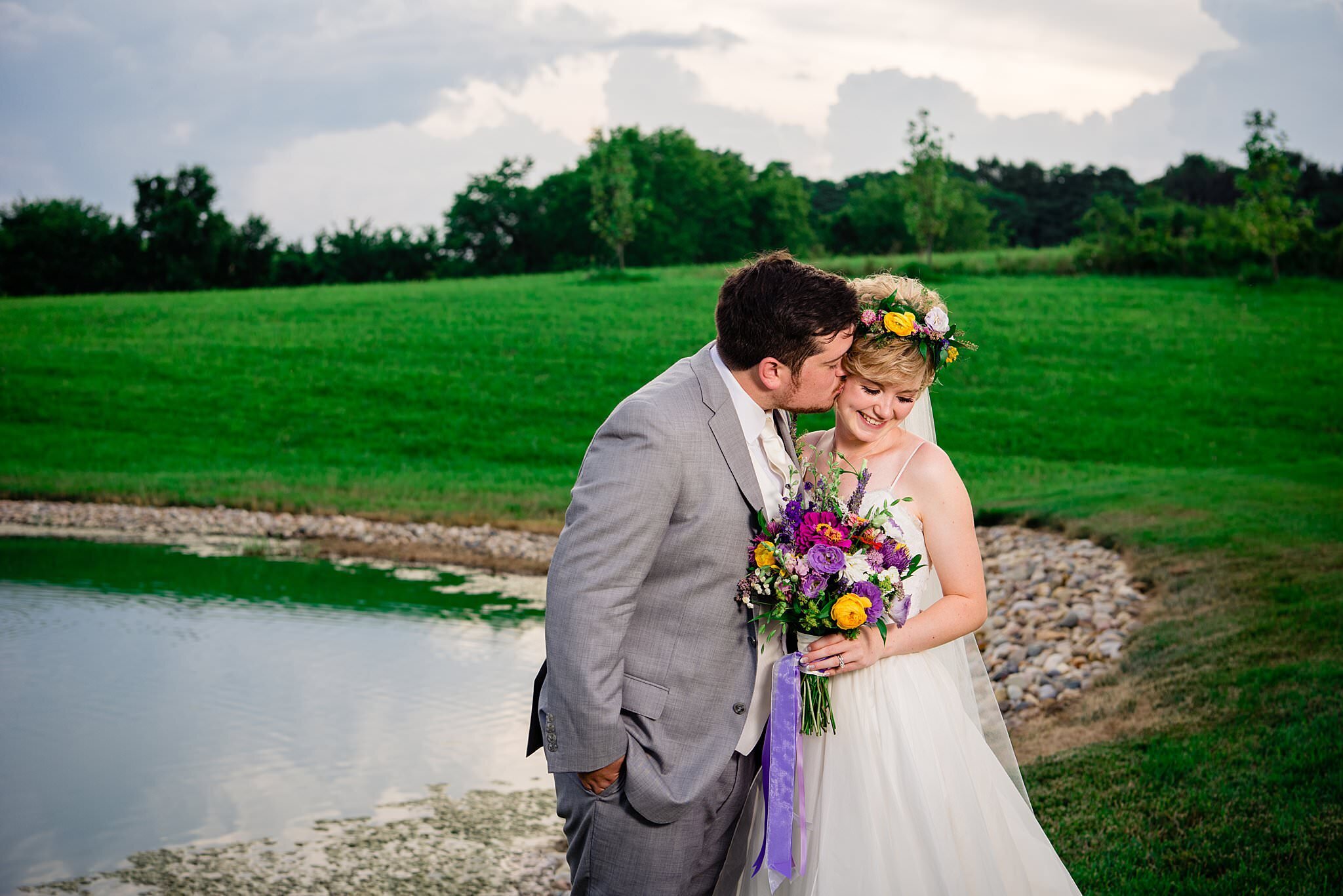 Bride wearing vibrant flower crown and bouquet standing beside a pond at White Dove Barn