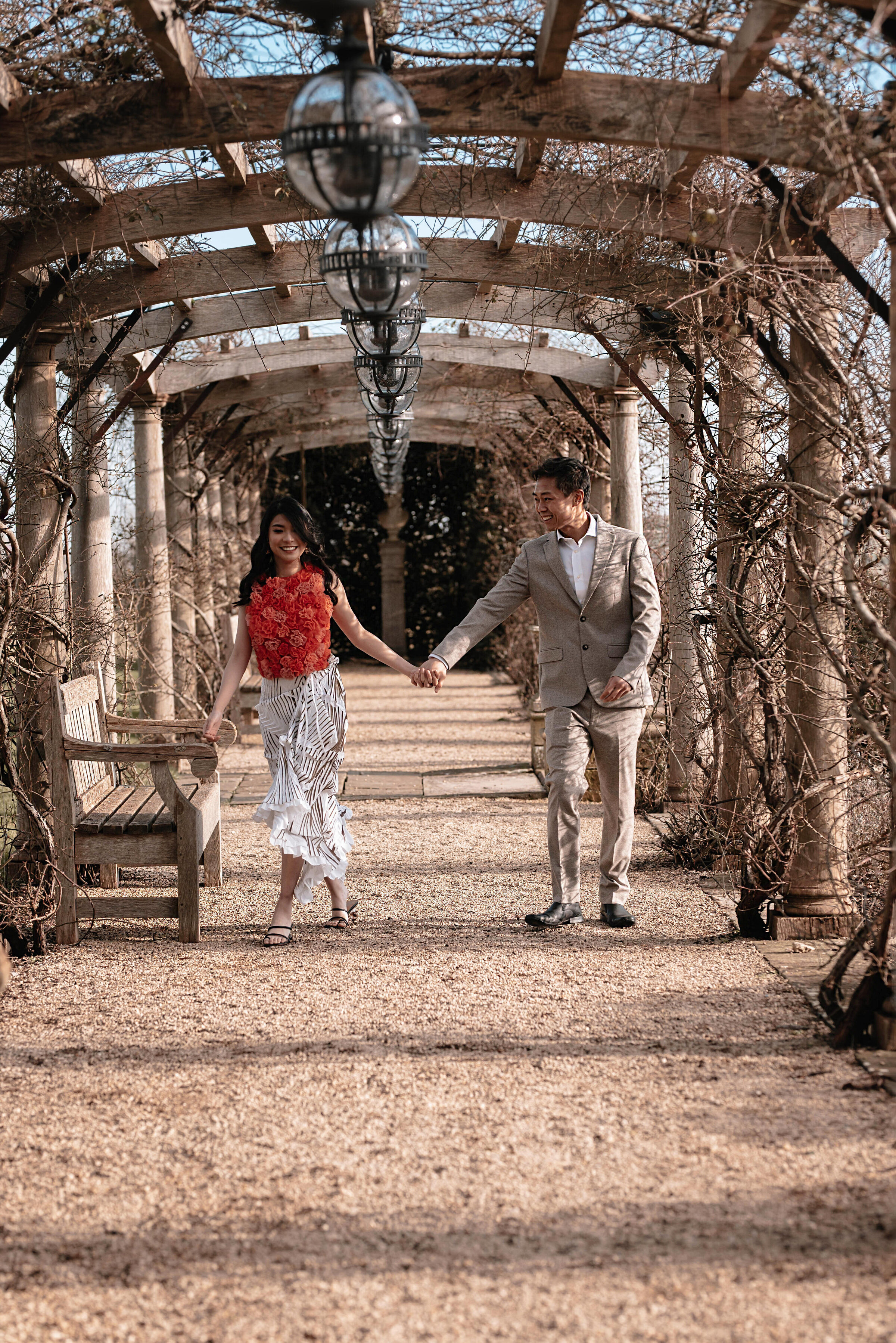 Man and woman laughing and walking hand in hand towards the camera, underneath a pergola entwined with vines