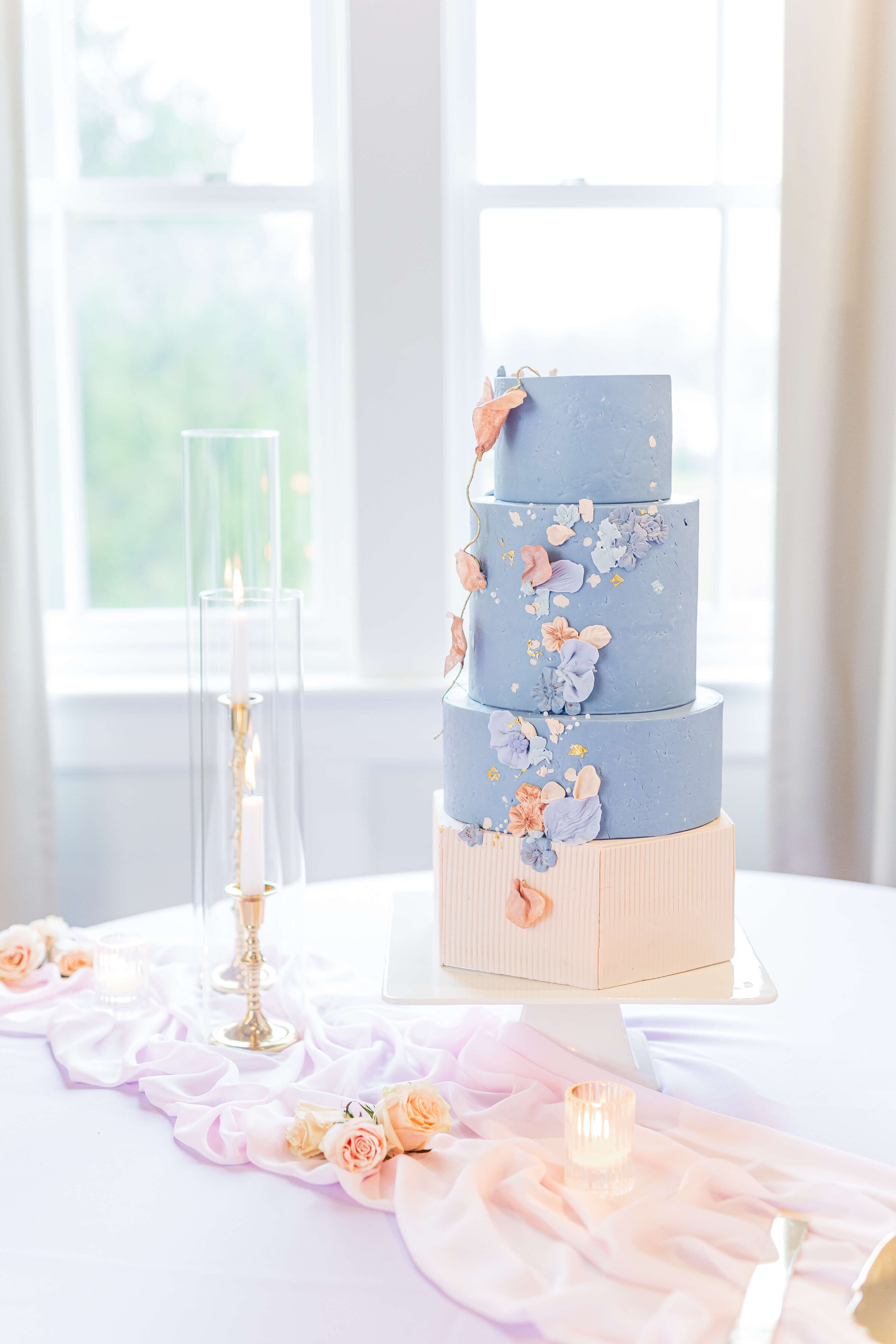 A wedding cake with a light pink bottom tier and two blue tiers on top of it with smaller flowers on the cake. A candlestick sits on the table next to the cake. Taken by a Dayton wedding photographer