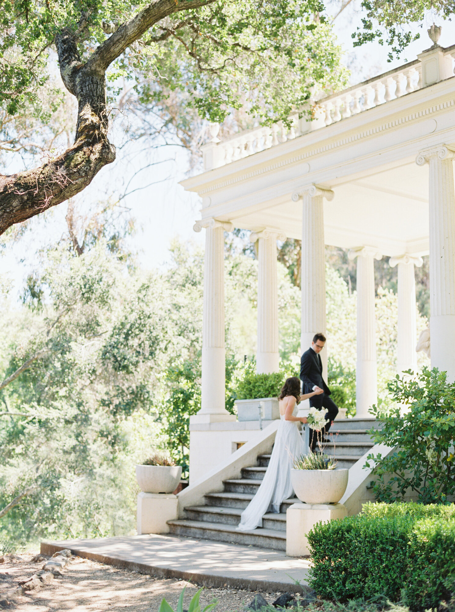 groom helping bride up the stairs of historic white pillar in garden