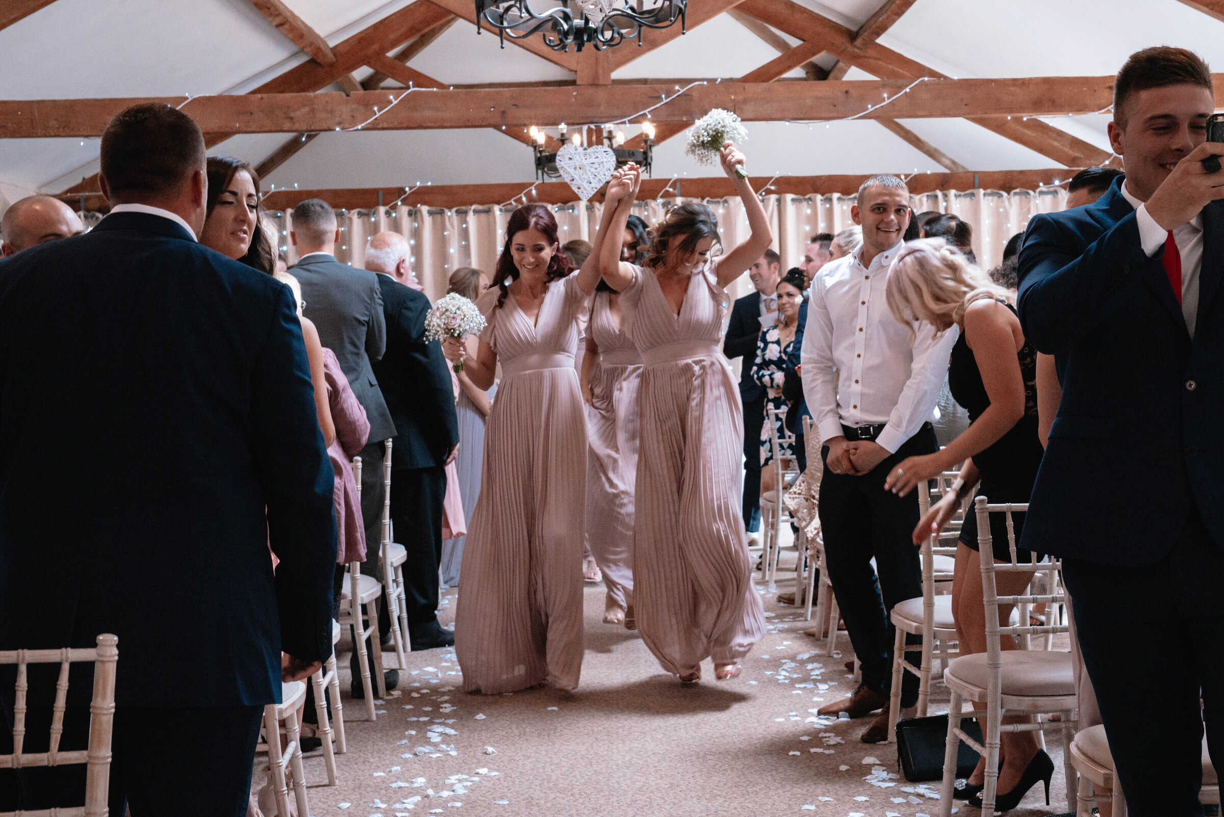 Bridesmaids dancing down the aisle holding their bouquets in the air whilst wedding guests clap