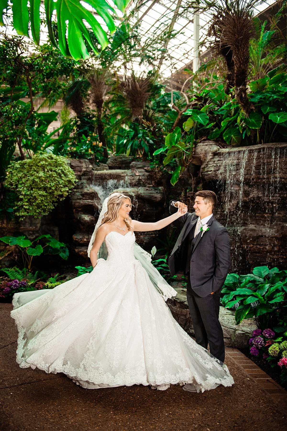 Groom spinning his bride who is wearing a Cinderella ballgown next to one of the atrium waterfalls inside the Gaylord Opryland Hotel
