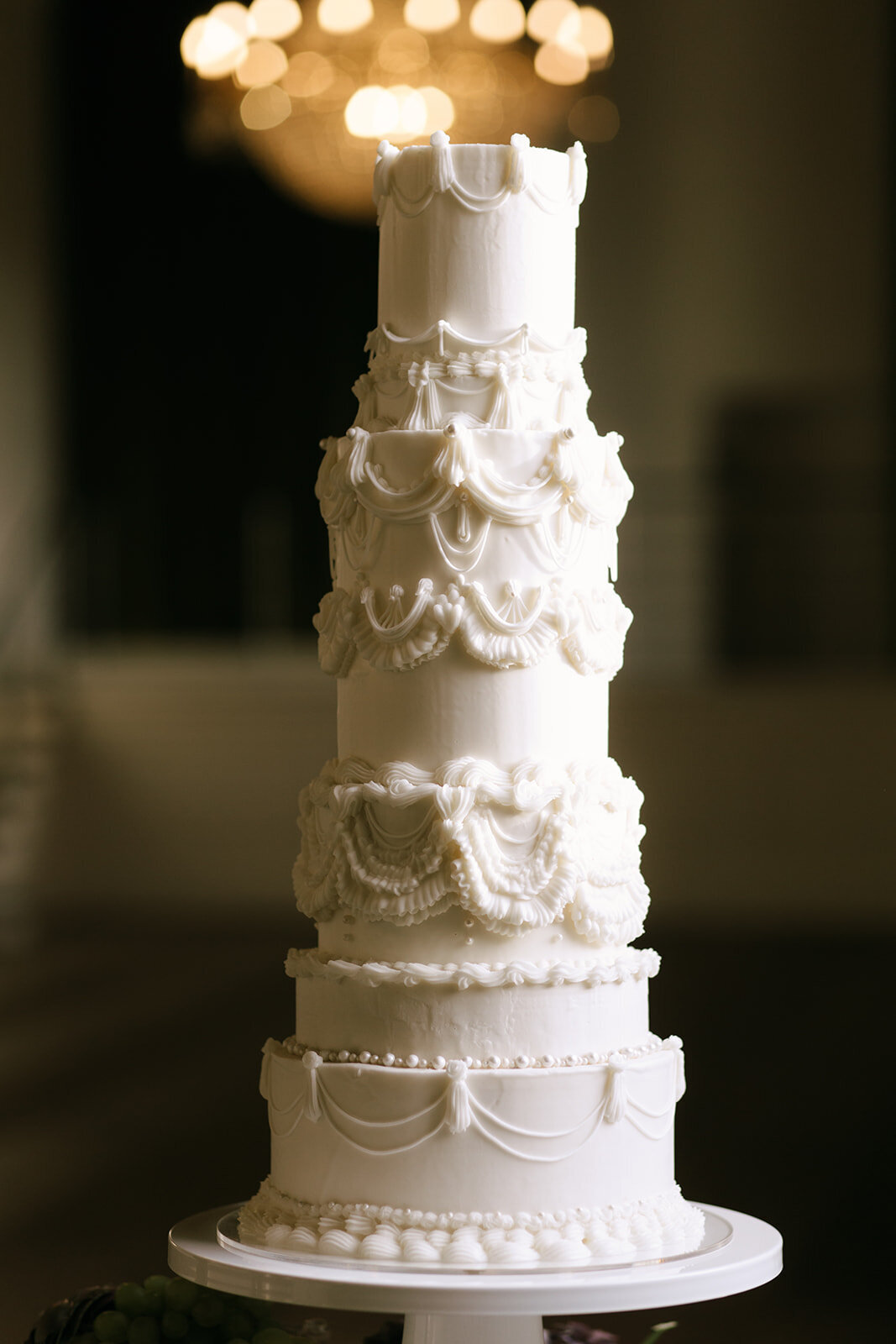 A tall white wedding cake with intricate draping details, inspired by luxury and timeless elegance, set against a softly lit background