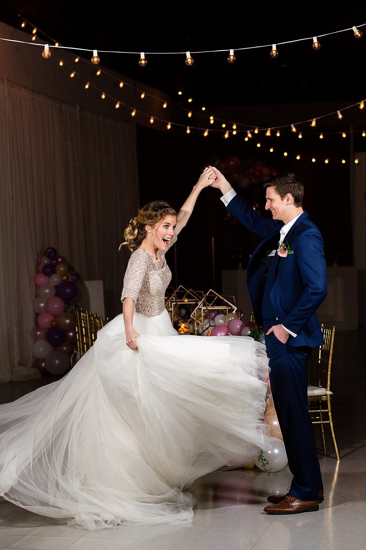 Bride spinning in her large tulle ballgown with her husband underneath indoor bistro lights