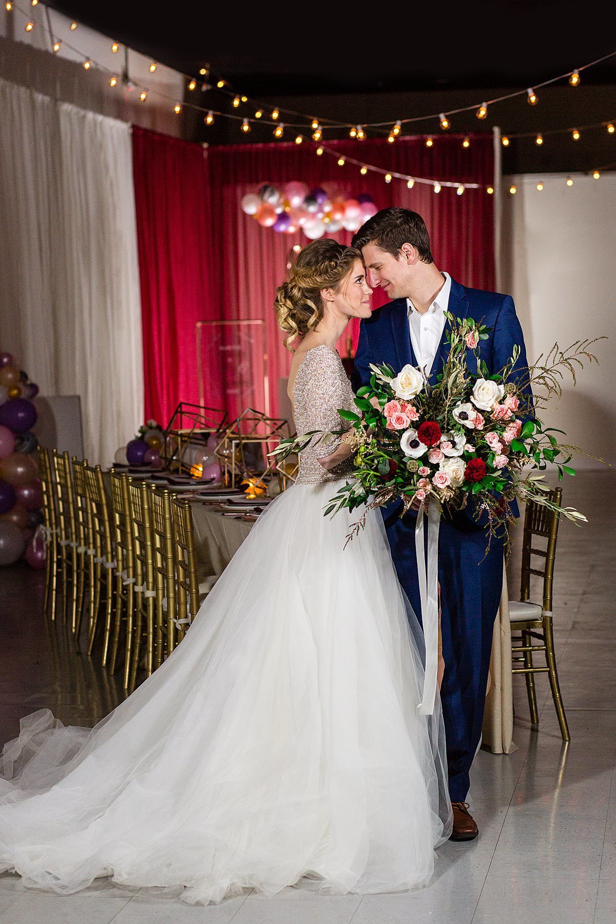Bride in sequin top with tulle skirt nose-to-nose with groom who's wearing a blue suit holding an oversized maroon and greenery bouquet