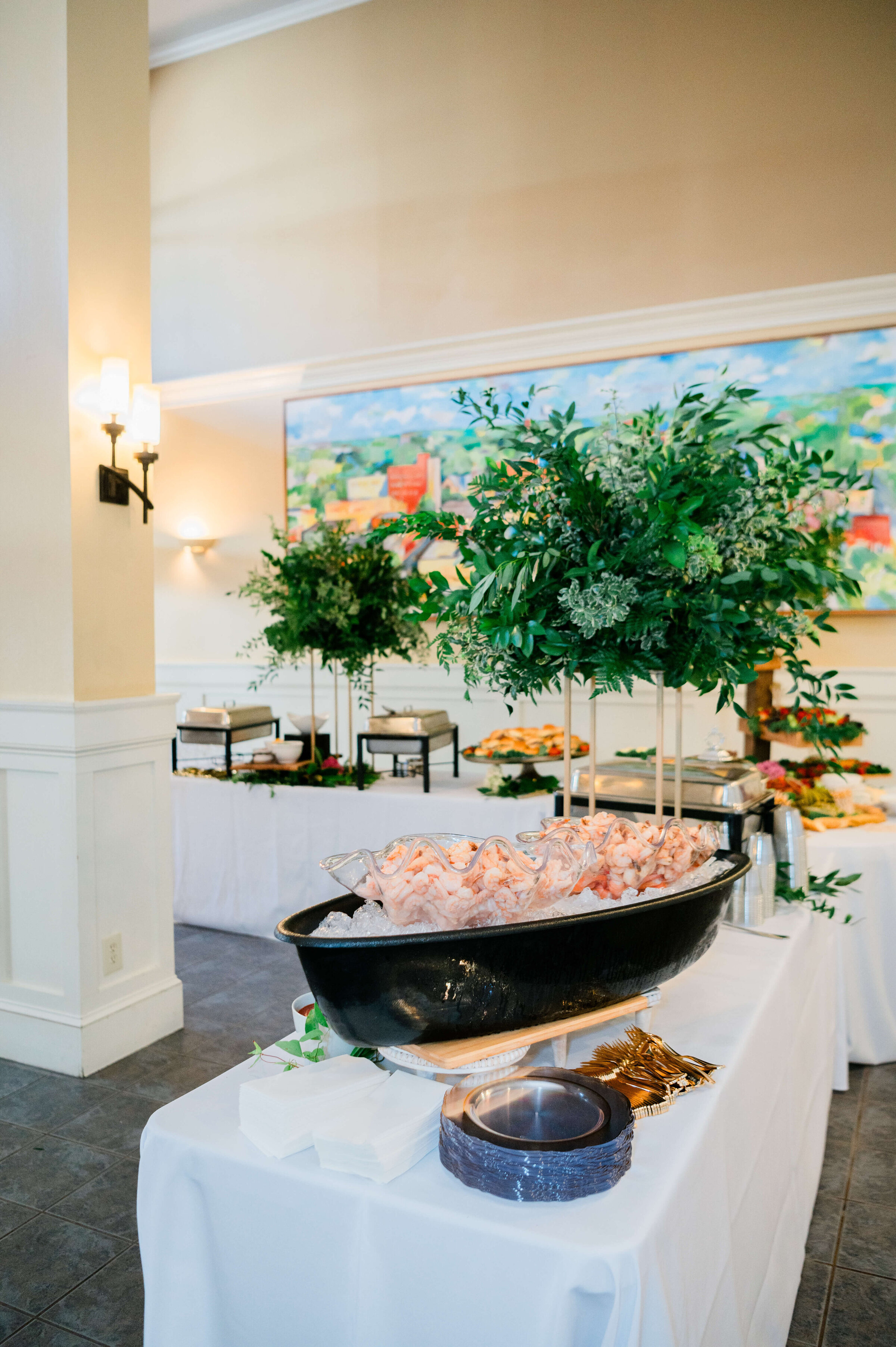 A buffet table at an event displays a large bowl filled with shrimp on ice, surrounded by plates, napkins, serving utensils, and decorative greenery, with more food and greenery in the background.