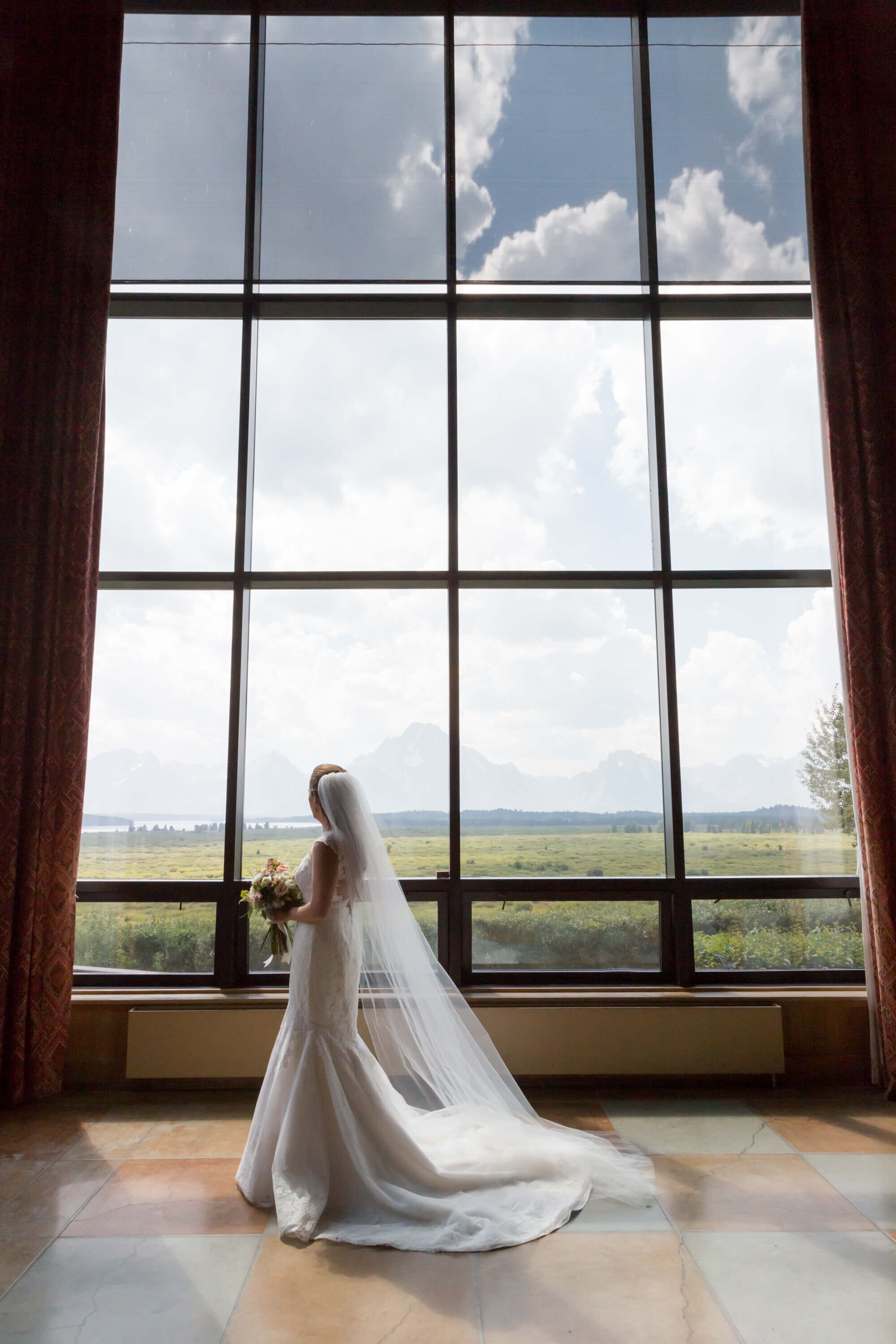 Bride by the Window at Jackson Lake Lodge – A bride in a white gown stands before the towering windows at Jackson Lake Lodge, gazing out at the Teton Range. Her veil cascades behind her as sunlight filters through the glass.