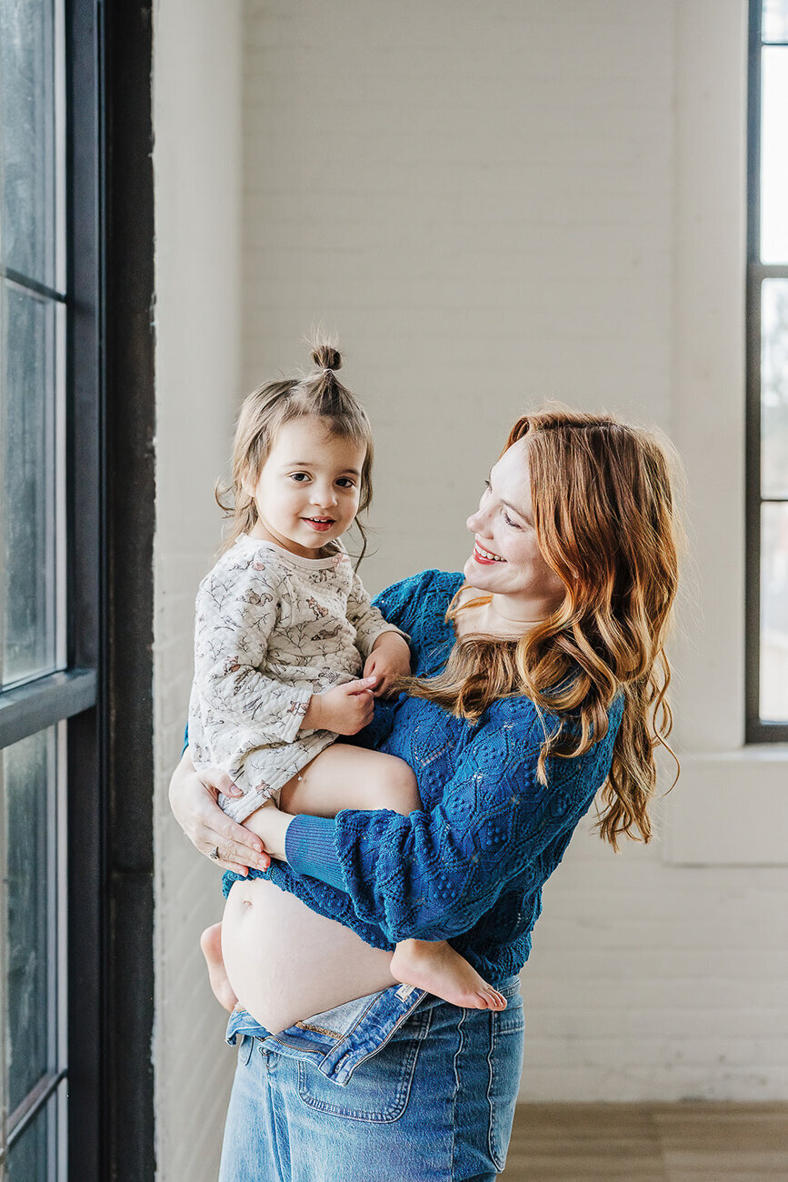 mom with bare belly carries toddler daughter on top of bump