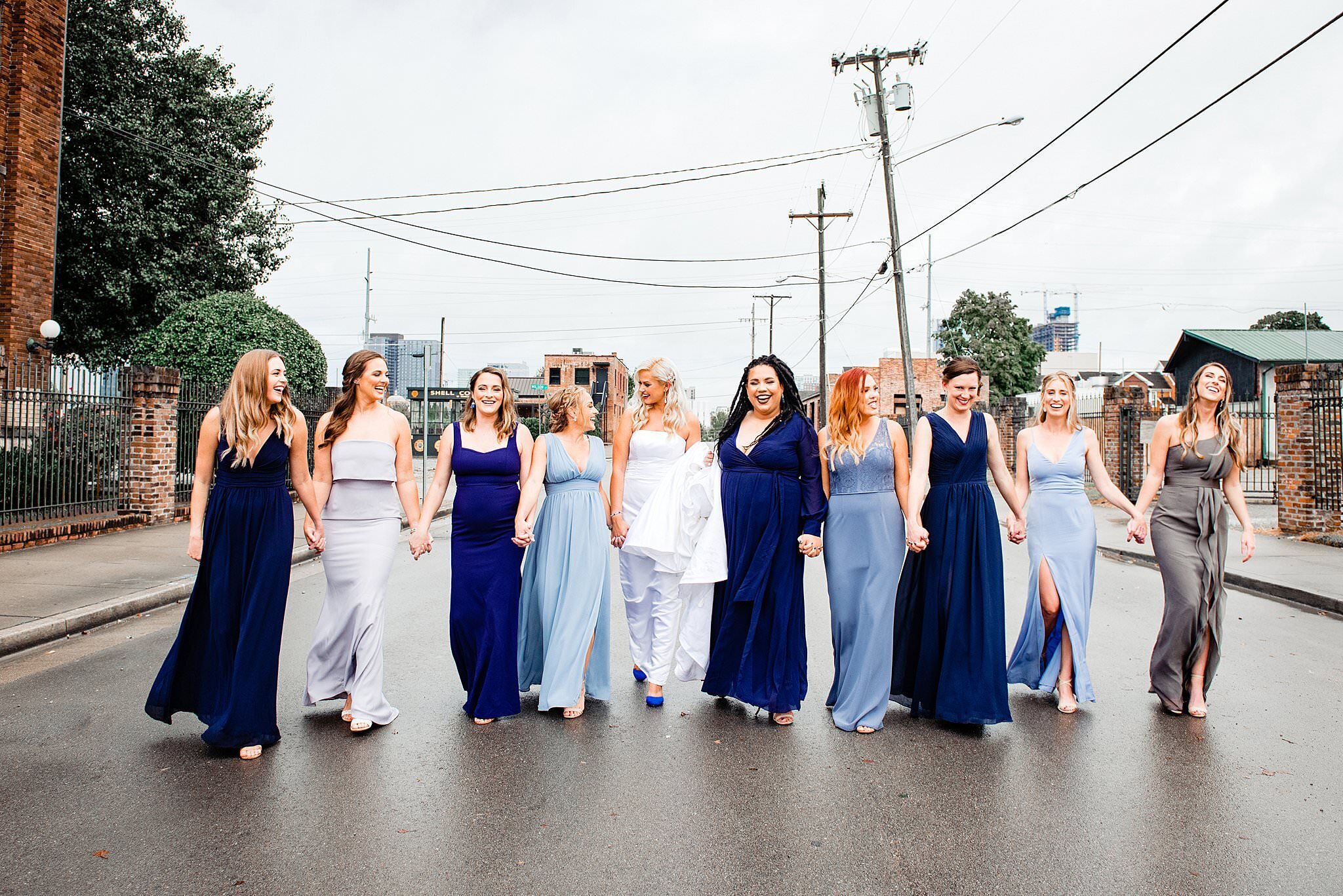 Bridesmaids wearing shades of blue from light to dark holding hands and strolling down an open roadway in Nashville near Marathon Village