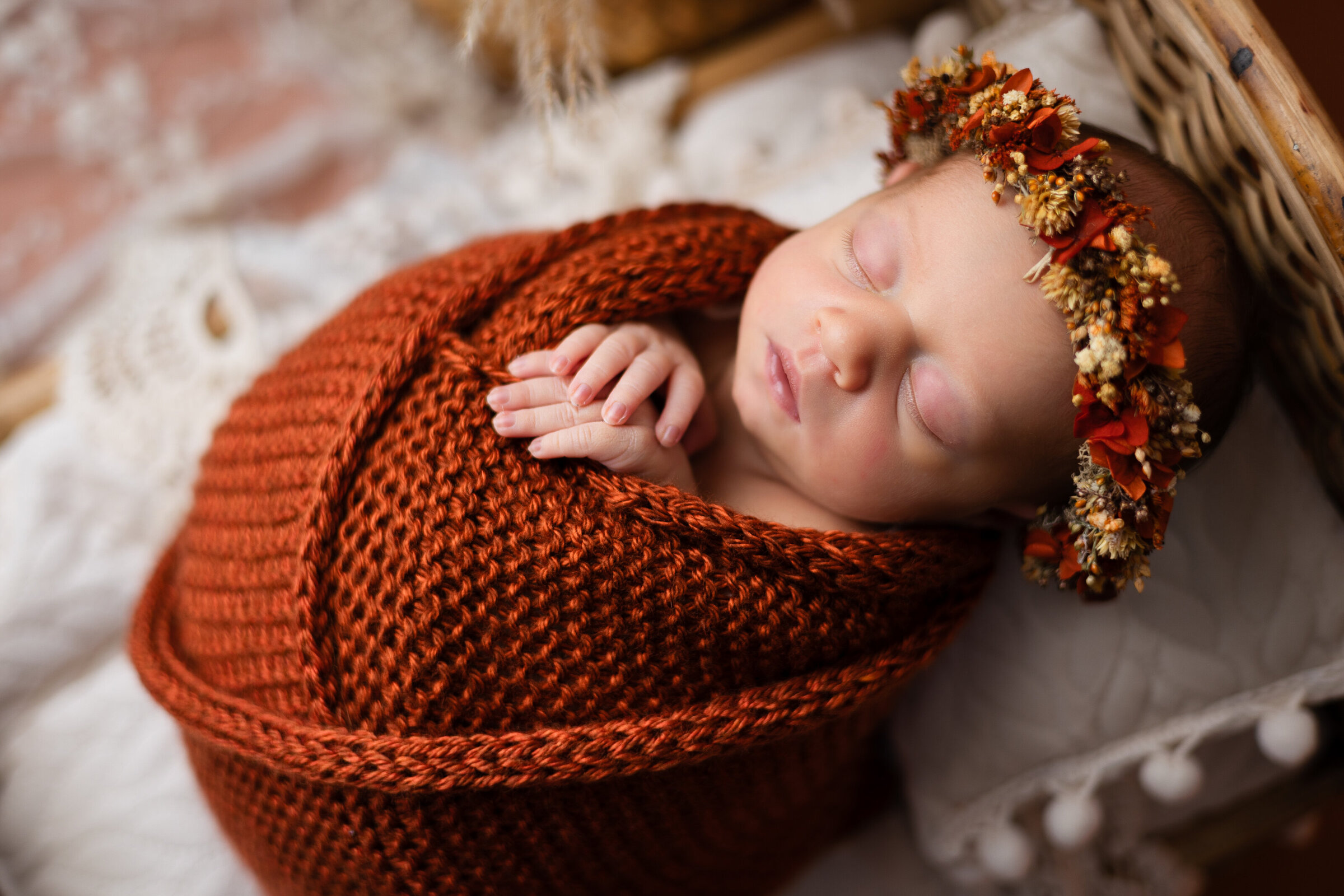 Newborn baby girl wrapped in rust orange with a matching floral halo , posed on a tiny bed