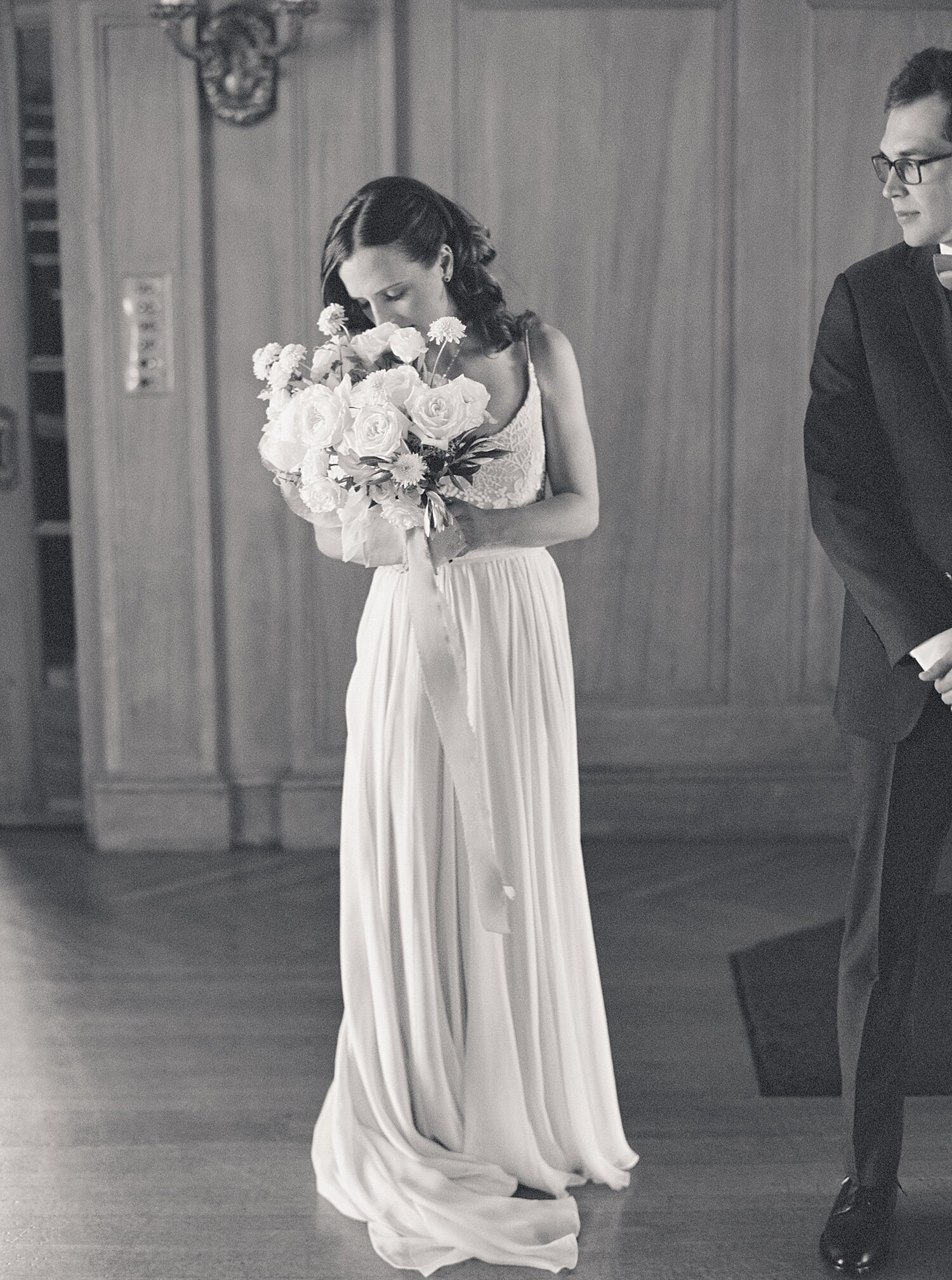 black and white film photo of bride in long chiffon gown smelling her rose bouquet