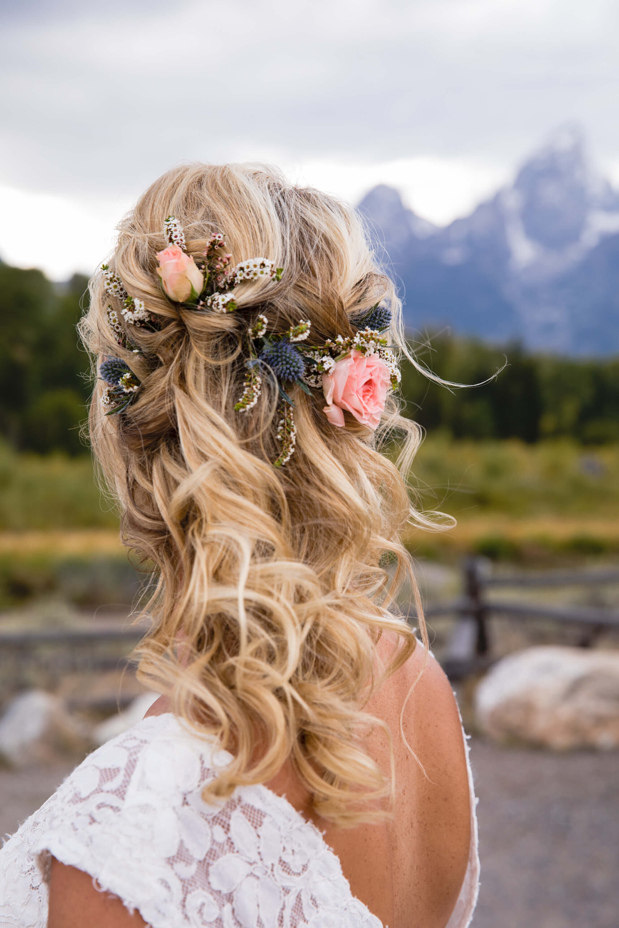A bride’s intricate bridal hairstyle is adorned with blush roses, white floral sprigs, and blue thistles. Loose waves cascade down her back as she stands in Grand Teton National Park, Wyoming, with mountain peaks in the background. Her lace wedding dress complements the natural elegance of the setting.