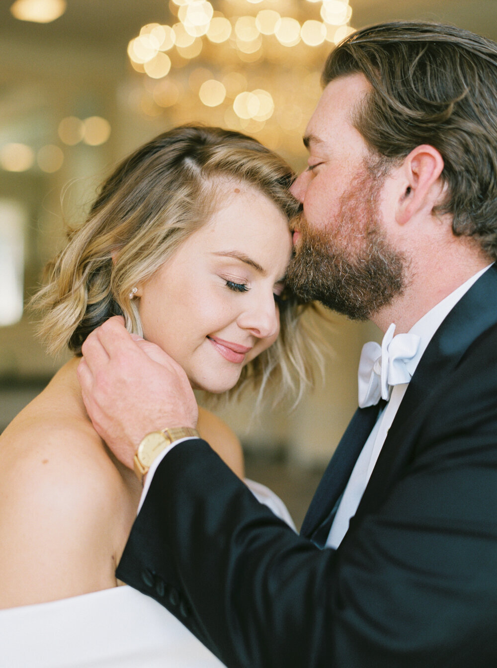 groom kisses bride on the forehead with golden bokeh in background on film