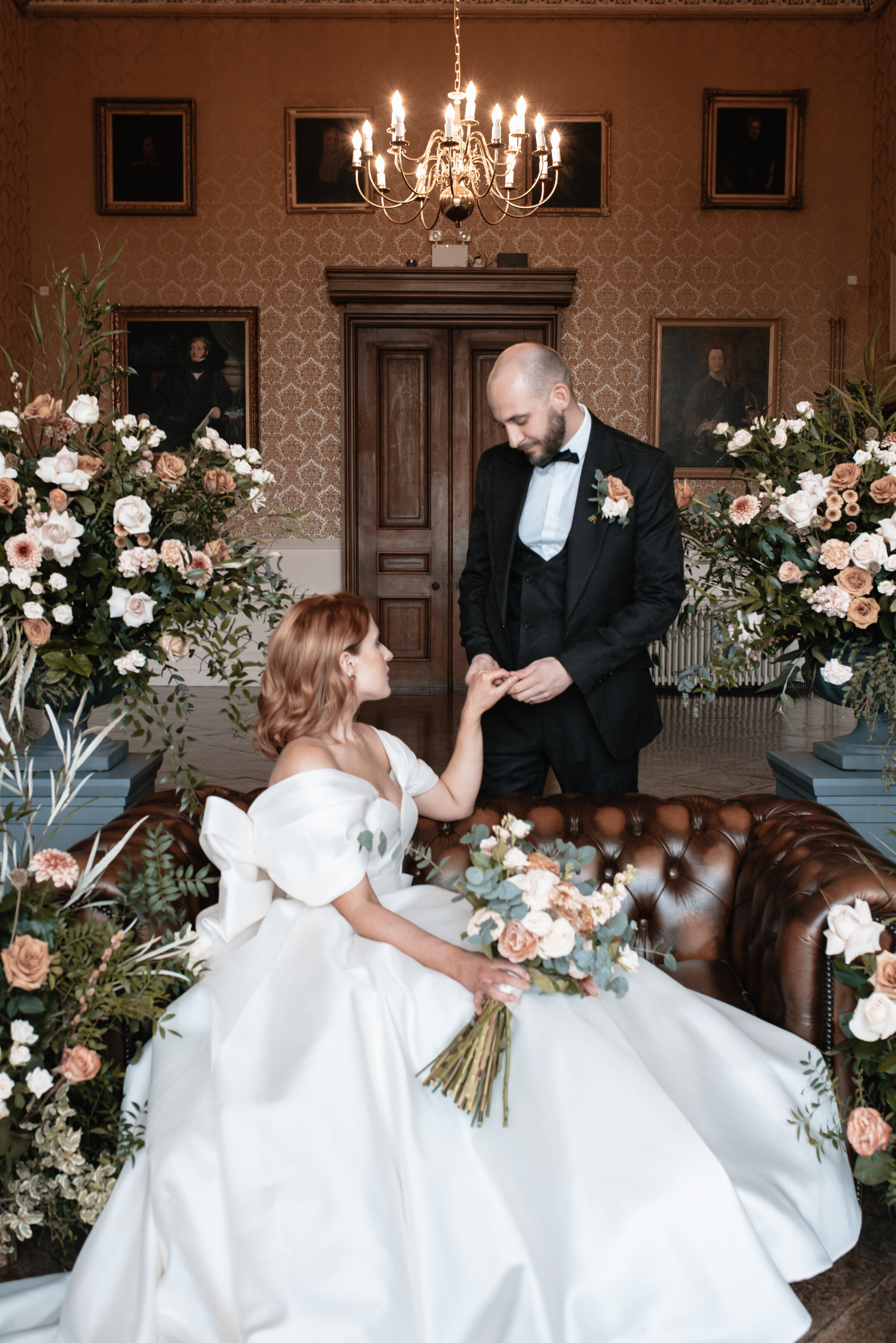 Bride seated holding her standing Grooms hand surrounded by beautiful floral arrangements in magnificent ballroom