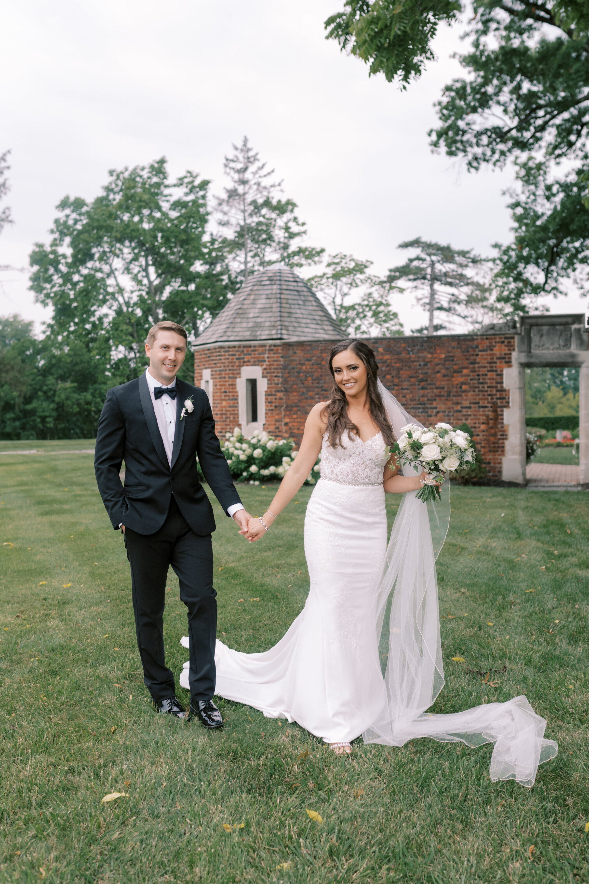 A bride and groom hold hands and walk alongside a brick exterior of Pinecroft Mansion in Cincinnati, Ohio