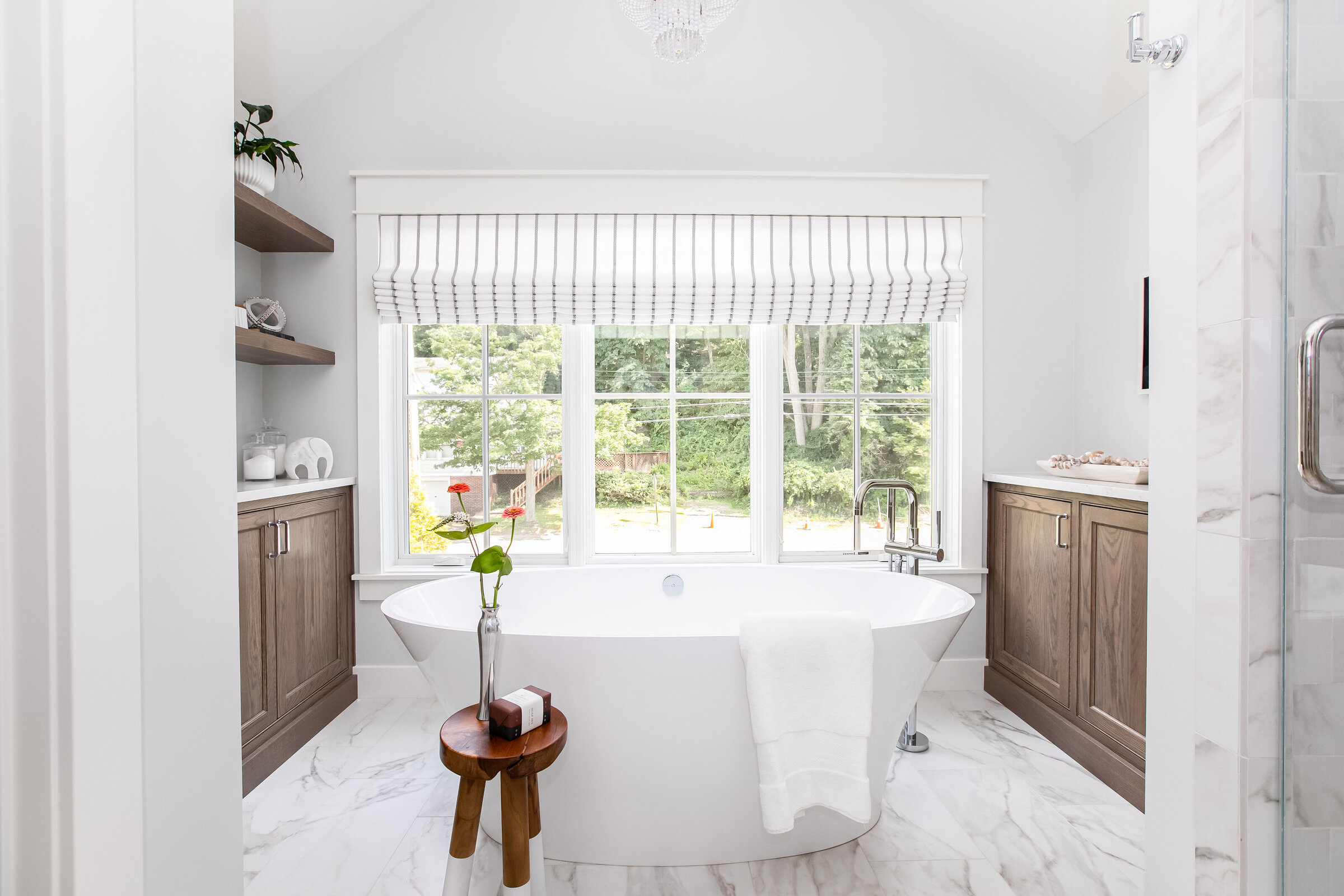 A serene bathroom featuring marble floors, a spacious soaking tub, and a charming stool adorned with flowers. Floor-to-ceiling windows offer a stunning view of lush green trees outside, creating a tranquil and elegant space for relaxation.