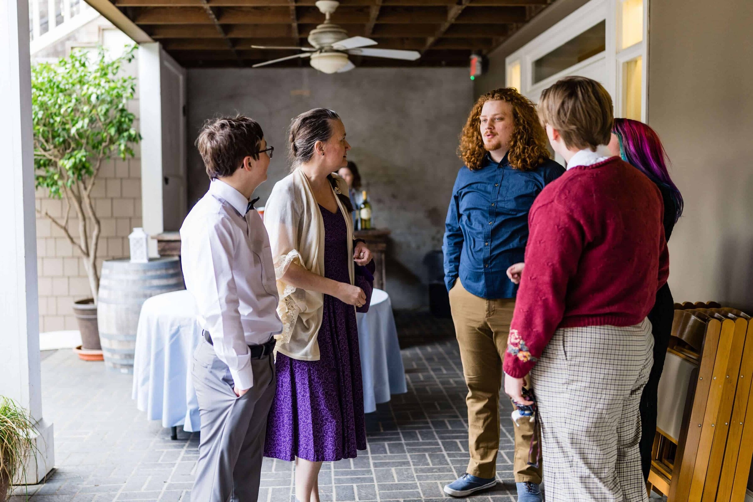 Five people stand in a casual group indoors, engaged in conversation at a private event. One person wears a purple dress, others wear shirts, pants, and sweaters. Tables, folding chairs, and a barrel are in the background.