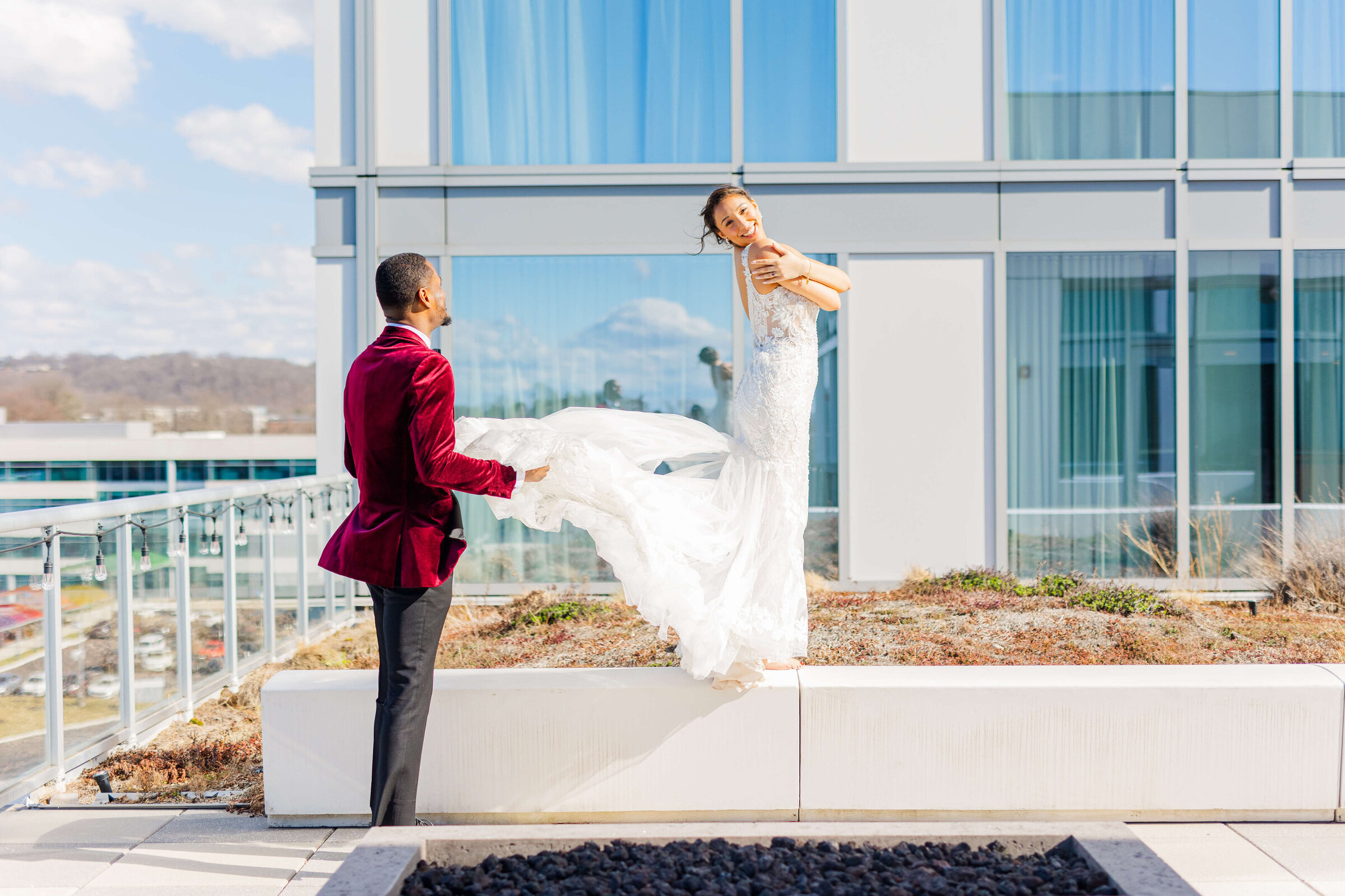 A groom in a maroon velvet tuxedo holds the wedding gown of a bride who is standing on a stair above him on top of a building. Taken by Dayton wedding photographer.