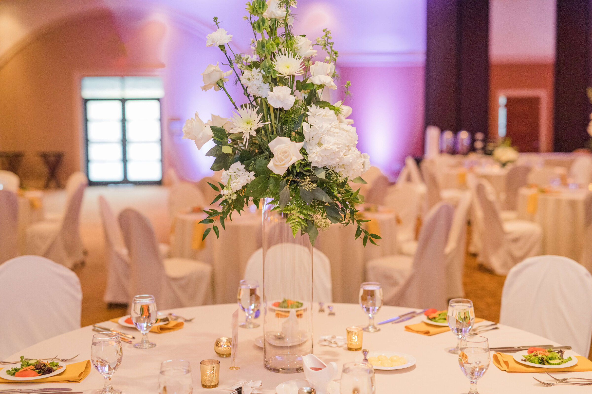 A photograph of a table at a wedding reception that has yellow napkins and tall flowers with purple lights in the background