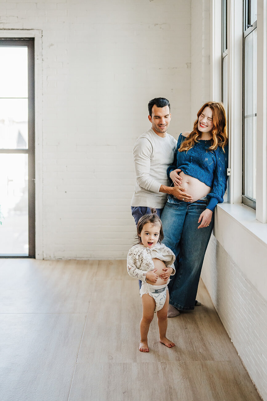 toddler girl shows belly in studio for maternity photoshoot