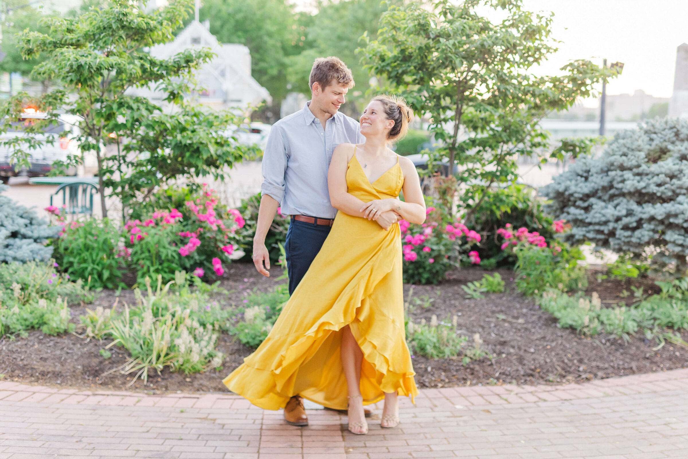 A man in a blue shirt and navy pants, twirls his fiance, who is wearing a bright yellow dress. They are standing at a park in Dayton ohio with lots of green trees and pink flowers.