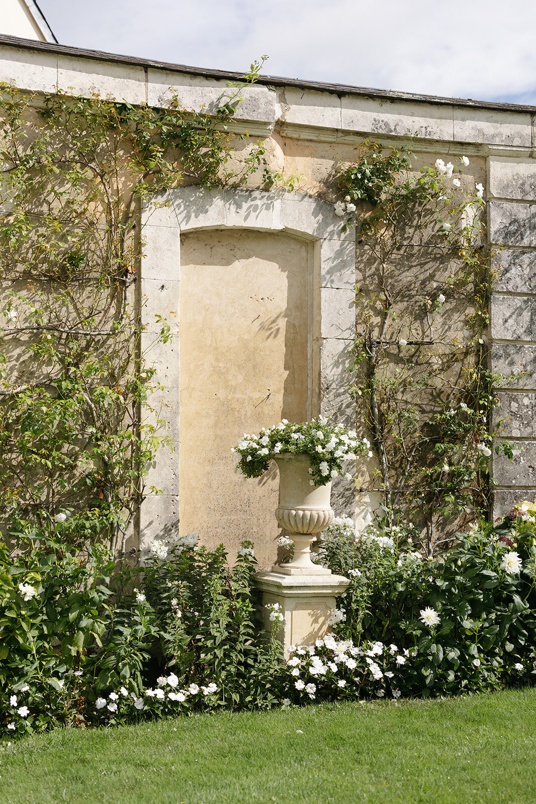 A garden wall at a French chateau featuring climbing greenery, white florals, and a decorative urn for a classic and elegant setting.