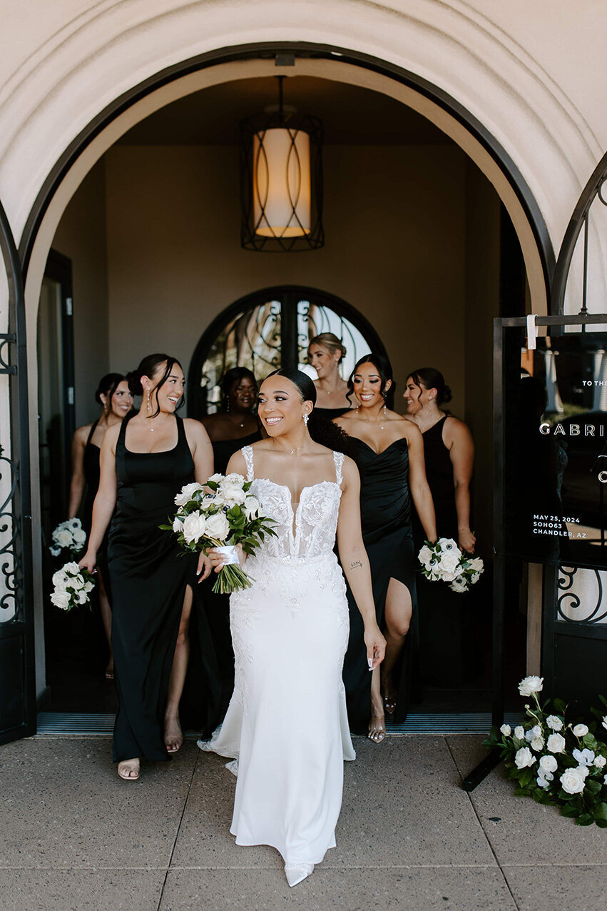 A radiant bride leads her bridesmaids out of the grand arched doorway at SoHo63, a Chandler wedding and event venue. Holding a white bouquet and smiling brightly, she walks arm-in-arm with her bridal party dressed in chic black gowns, capturing the confident and stylish energy of the day.