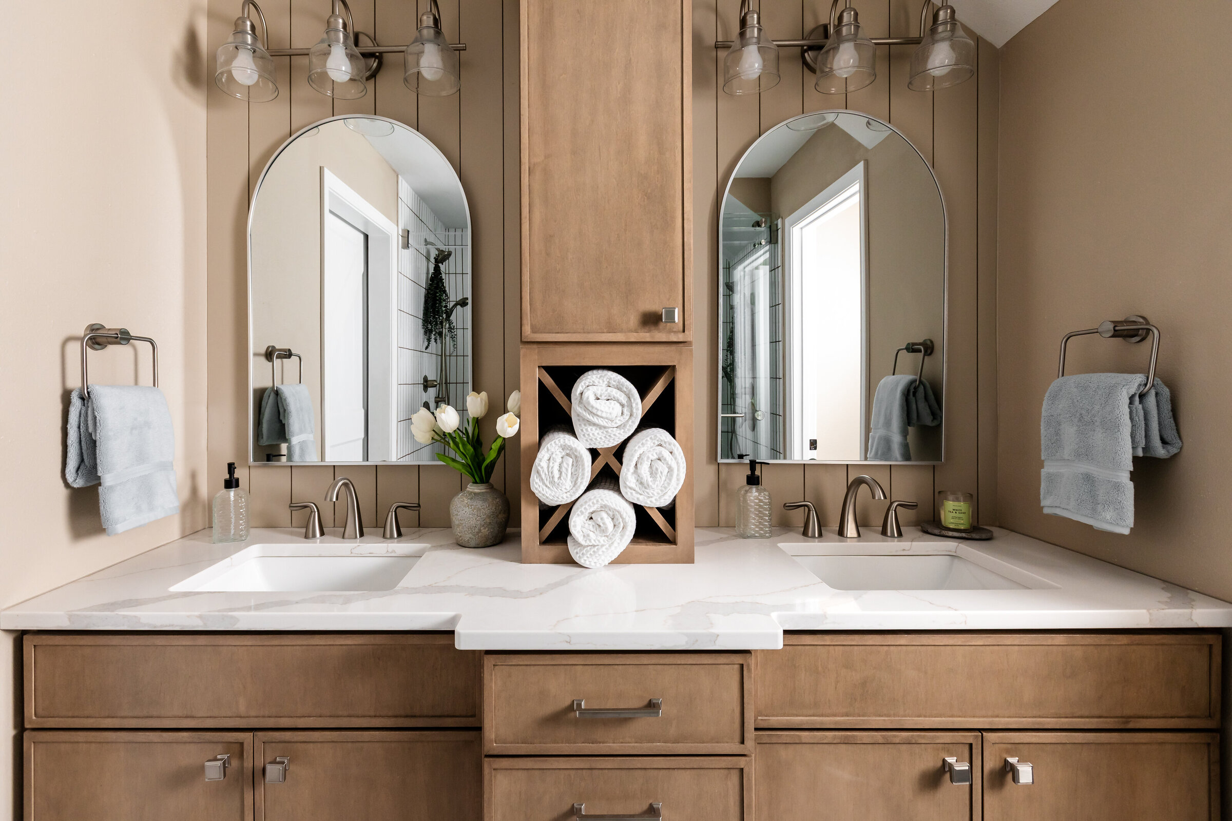 Large double bowl vanity in stained maple with tower storage between two sinks. Brushed nickel hardware and arched mirrors with towel rings on both sides.