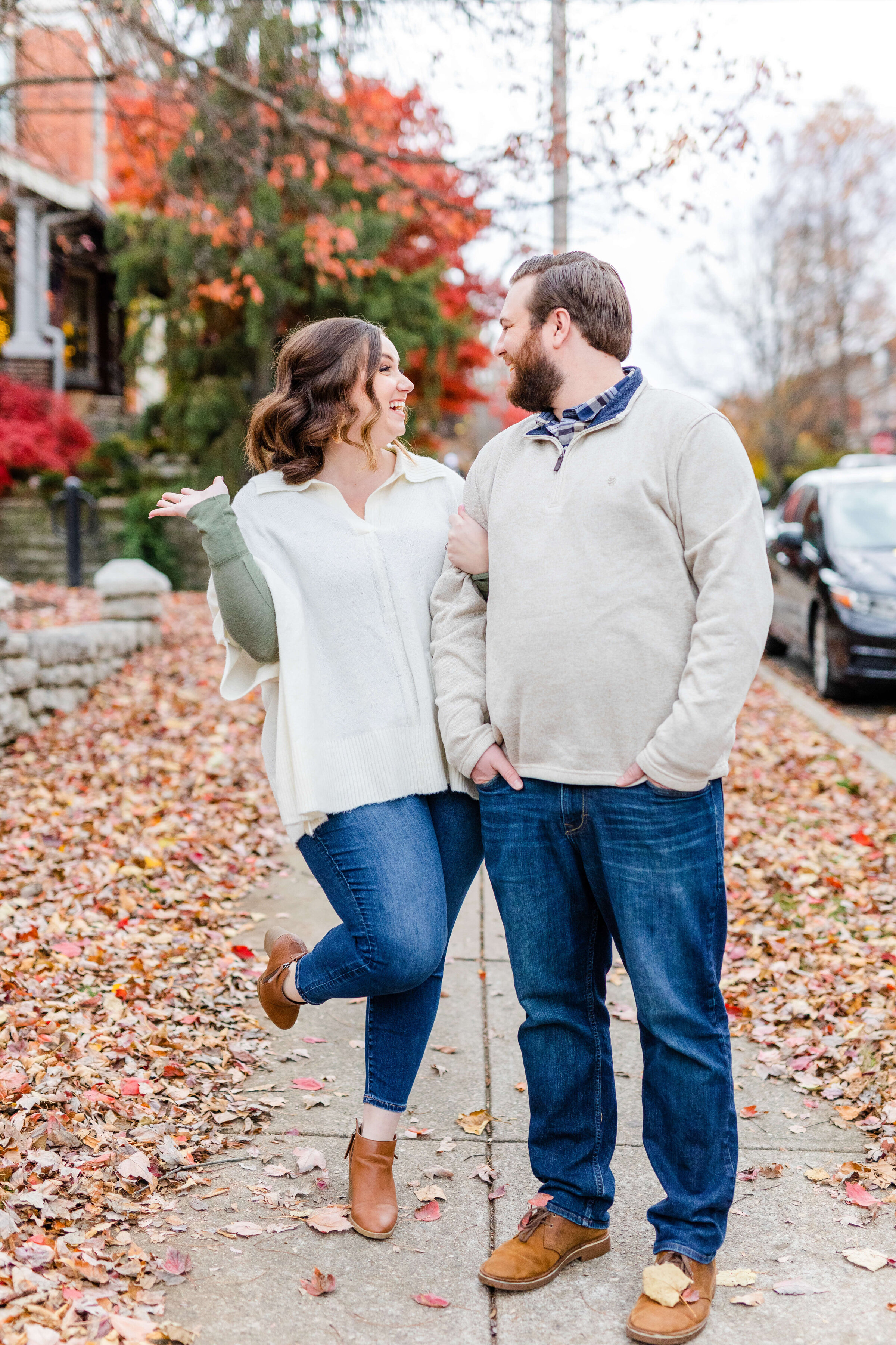 A man and woman in jeans and ivory sweaters stand arm in arm next to one another as the fall leaves are all around them. The girl is happy and has her leg and arm in the air. Taken by a lexington wedding photographer