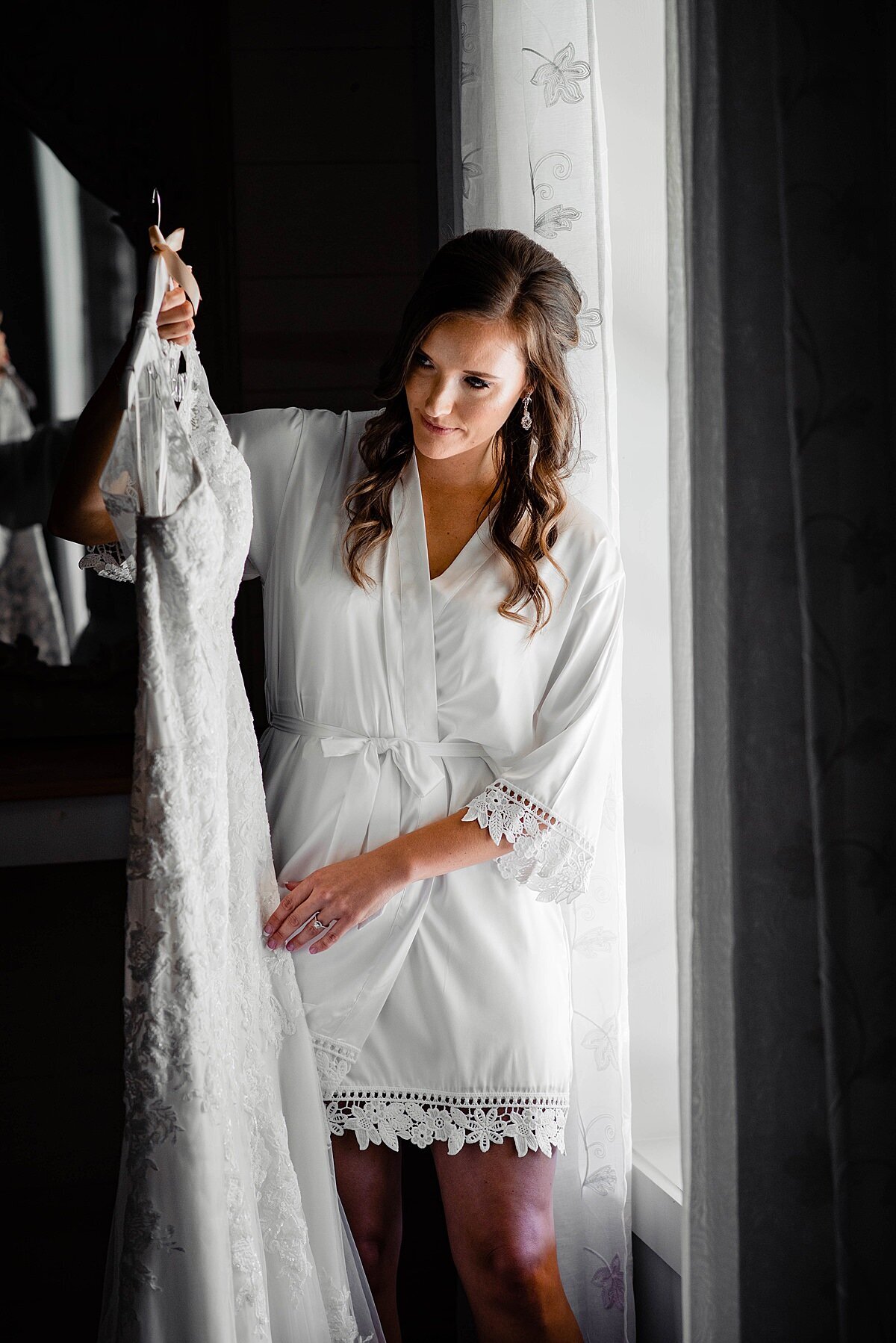 Bride standing beside the window in the bridal room at Cranford Hollow, her hair is curled and down and she has a white robe with lace trim on