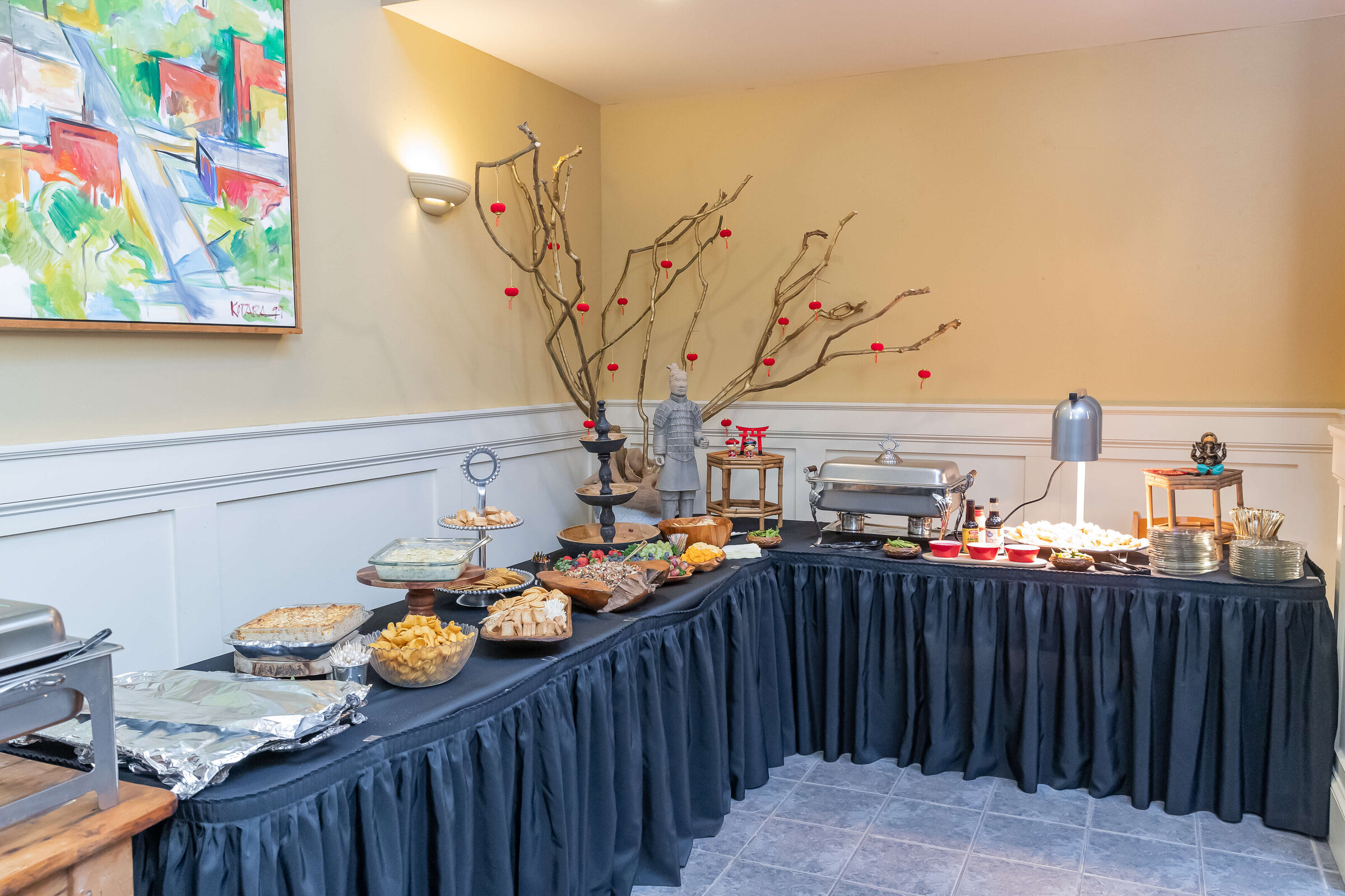 A buffet table in a corner, covered with a dark tablecloth and filled with various dishes, trays, and covered chafing dishes.