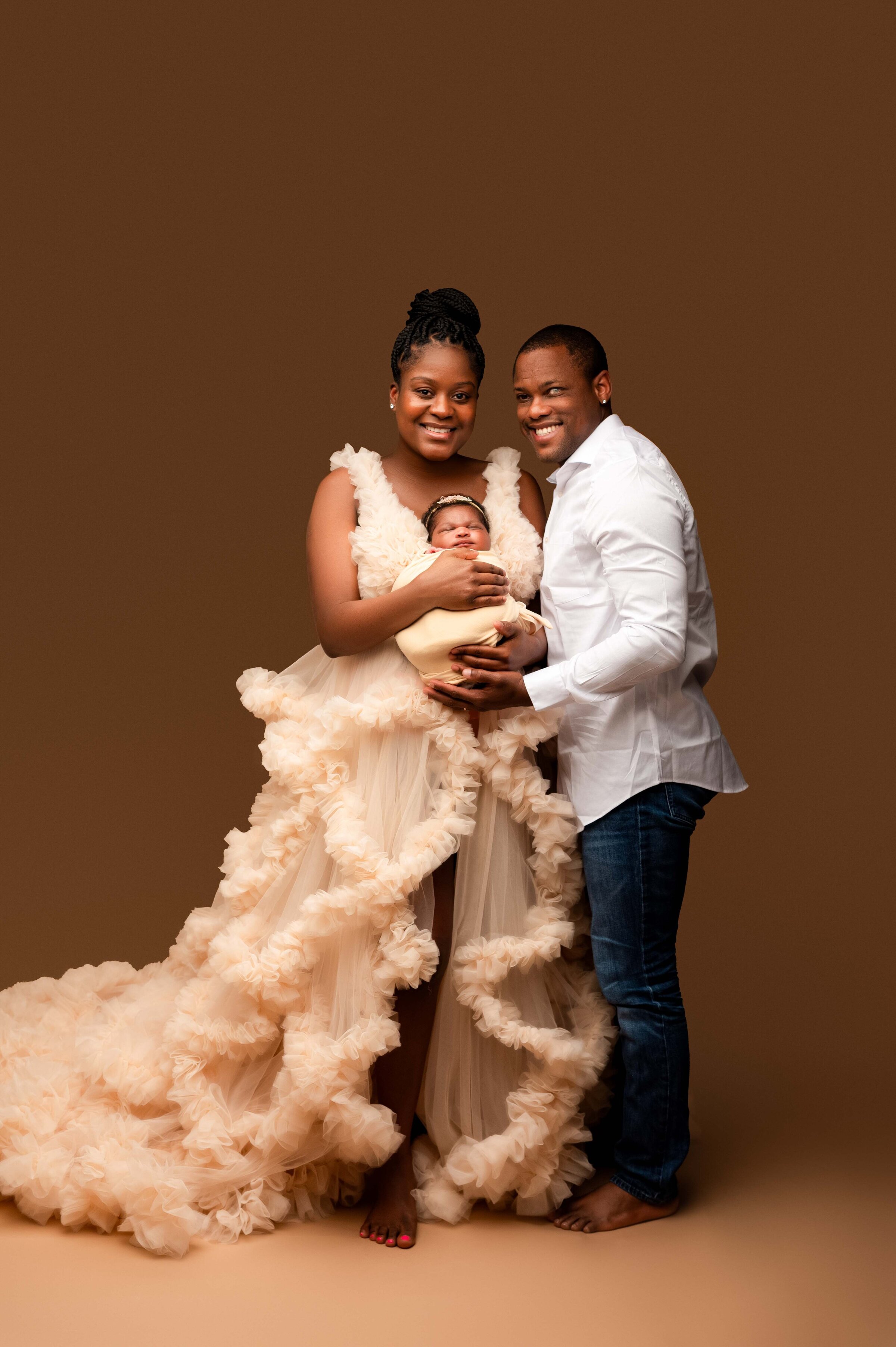 A couple stands smiling, holding a newborn baby wrapped in a blanket. The woman wears a voluminous beige gown, and the man is in a white shirt and jeans. Captured by a talented Waxhaw newborn photographer, they pose against a plain brown background.