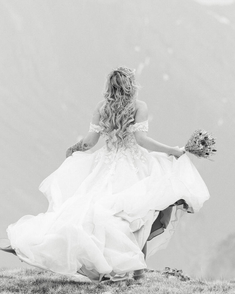 This stunning portrait captures a joyful bride as she playfully twirls her dress in the wind, moments after landing in the mountains via private helicopter. The flowing gown and breathtaking alpine setting create a magical, carefree moment amidst the rugged beauty of nature.