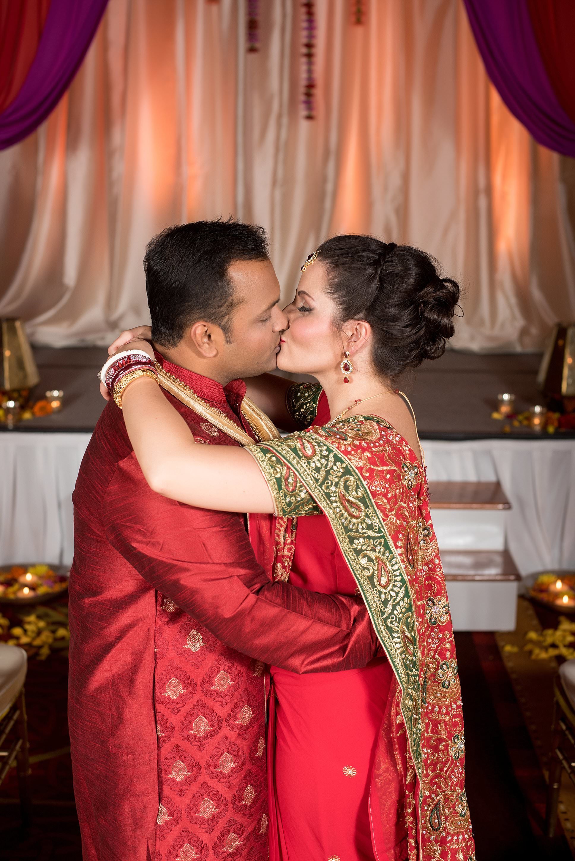 Bride and groom in their red wedding attire, lehenga kissing in front of their fuchsia and red mandap