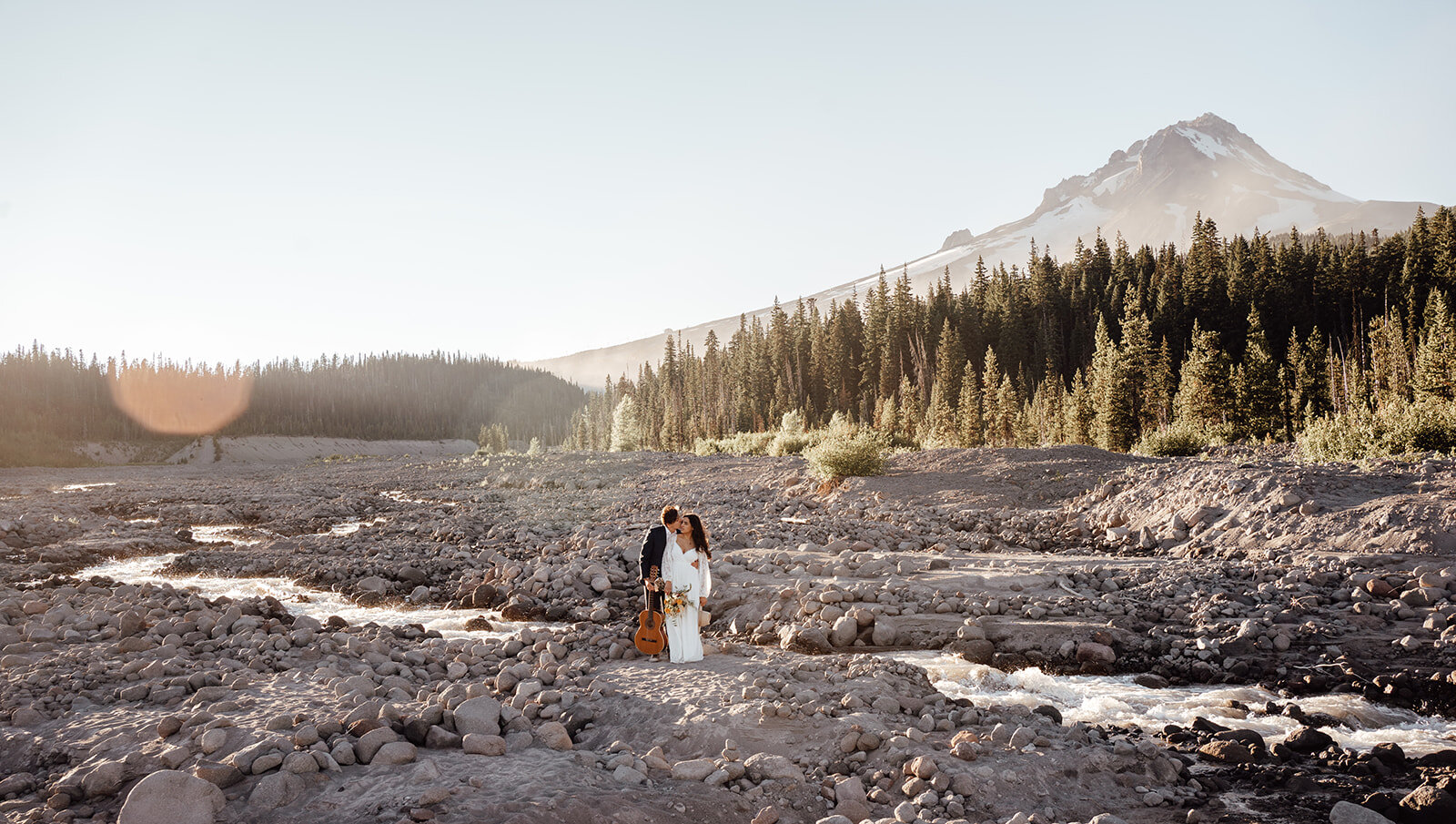 bride and groom standing in front of mt. hood. There is a river running through the back and they are embracing while holding a guitar.