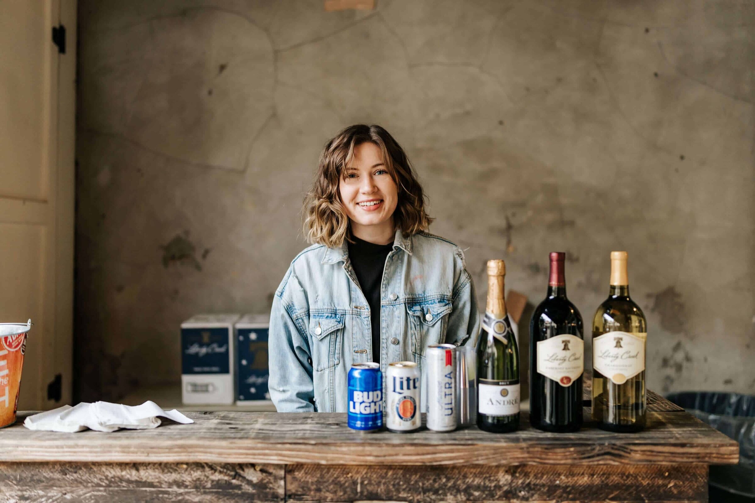 A woman with wavy hair wearing a denim jacket stands behind a wooden bar counter with various bottles displayed in front of her at a private event party.
